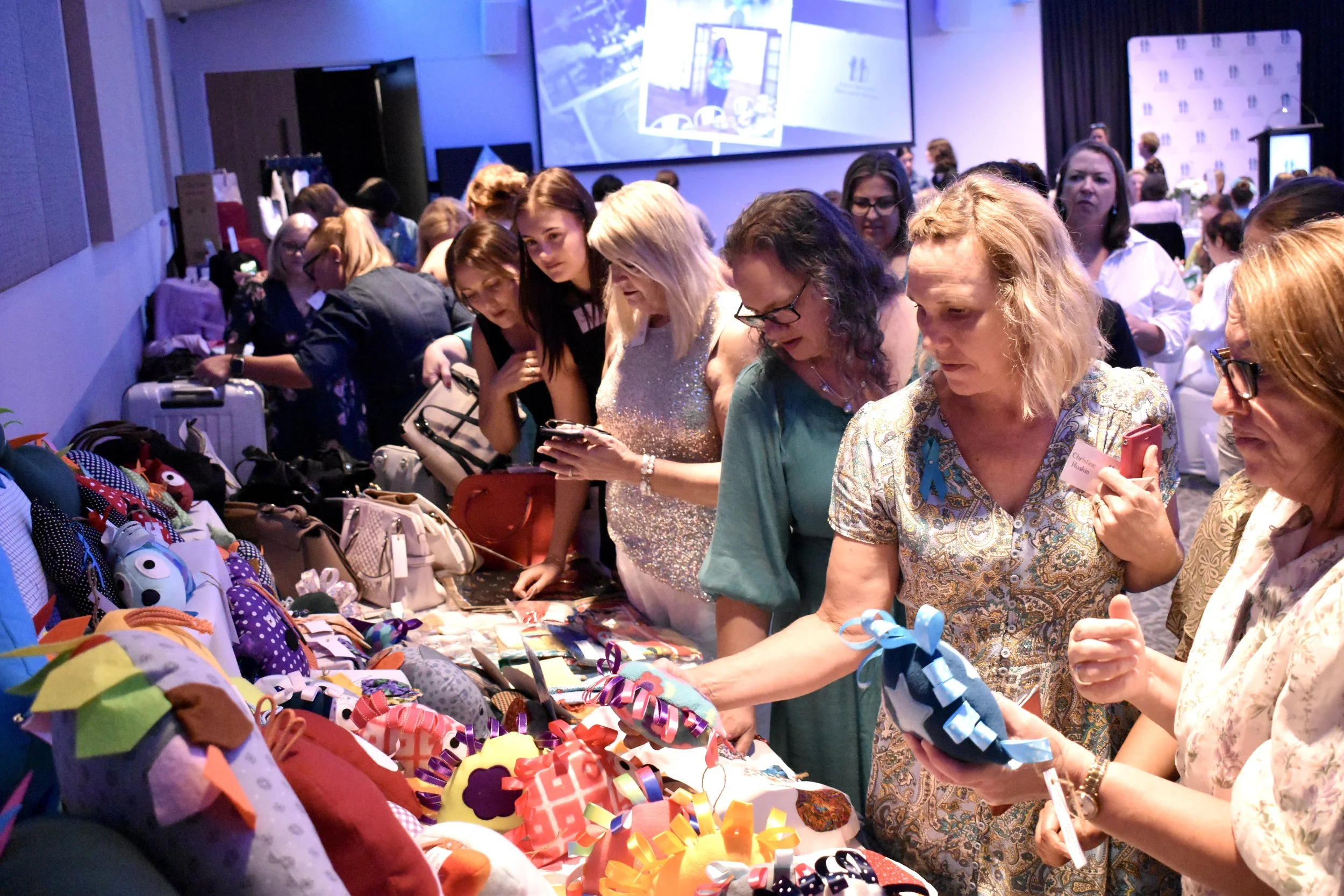 Women shopping at a market stall with colorful handmade crafts, bags, and ribbons.