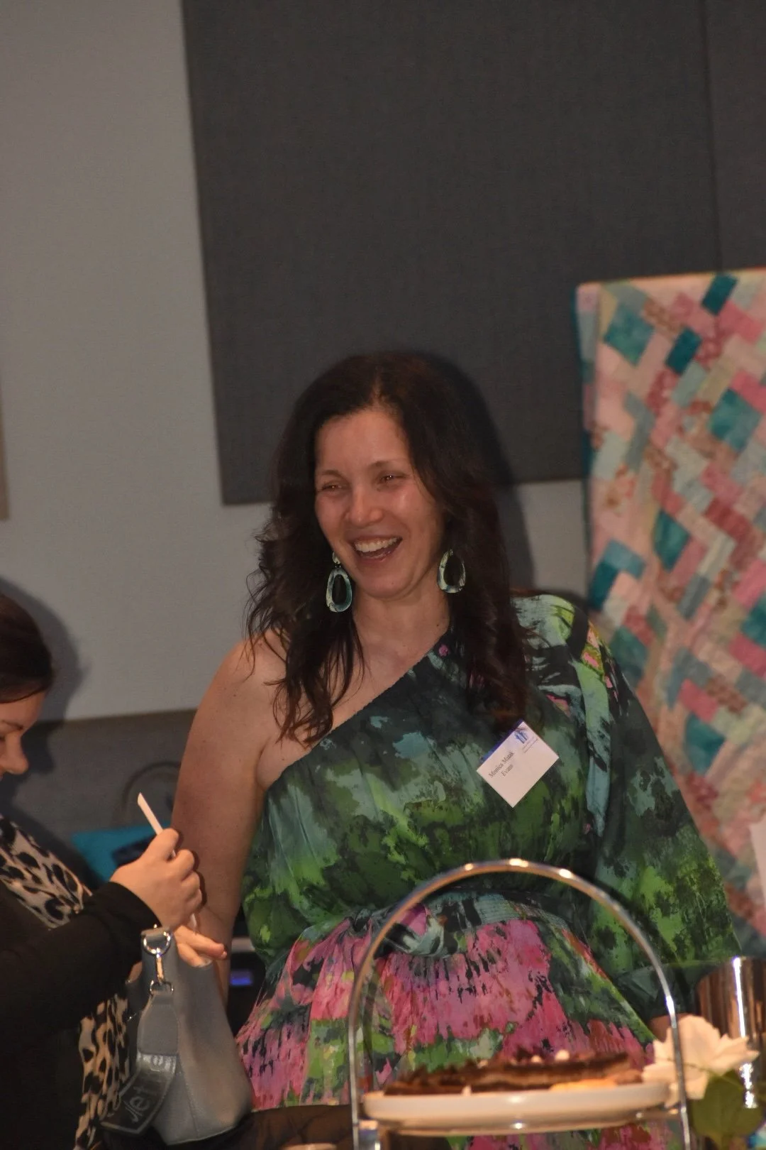 A woman with dark curly hair wearing large earrings and a colorful green and pink dress, smiling and laughing at a gathering.