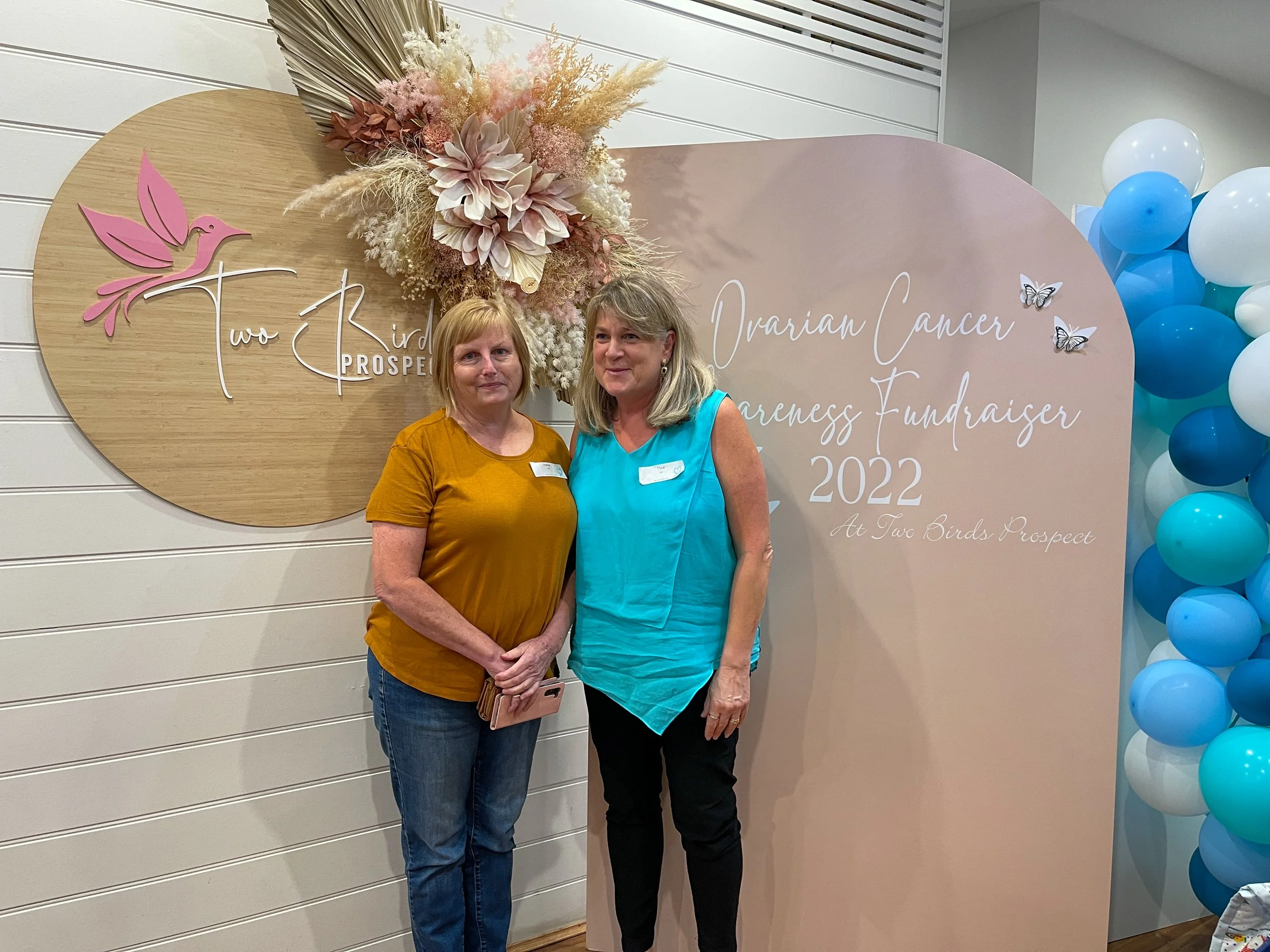 Two women standing in front of a sign for a ovarian cancer awareness fundraiser in 2022. The sign has a pink and white floral arrangement, butterfly decorations, and a logo with a pink bird. One woman is wearing a mustard yellow shirt and jeans; the 