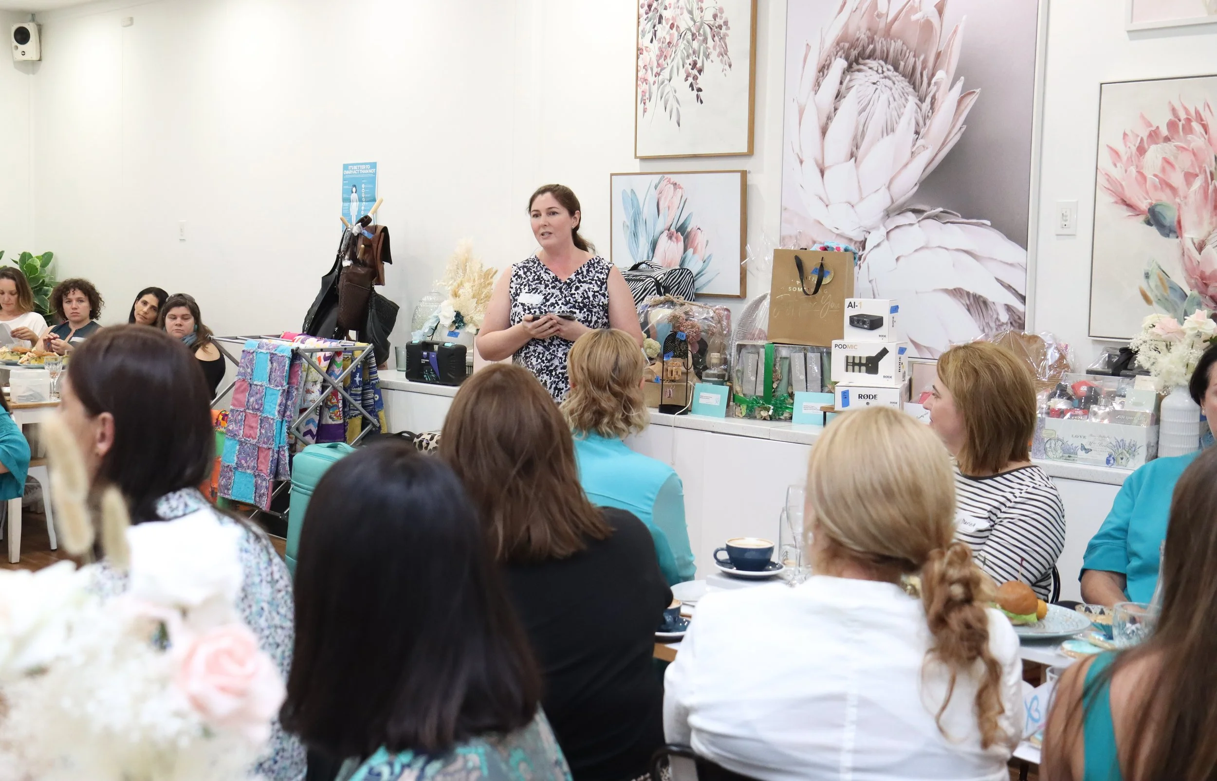 A woman standing and speaking to a group of women seated at tables, in a room decorated with large floral artwork on the walls, with various gift boxes and items on a counter behind her.