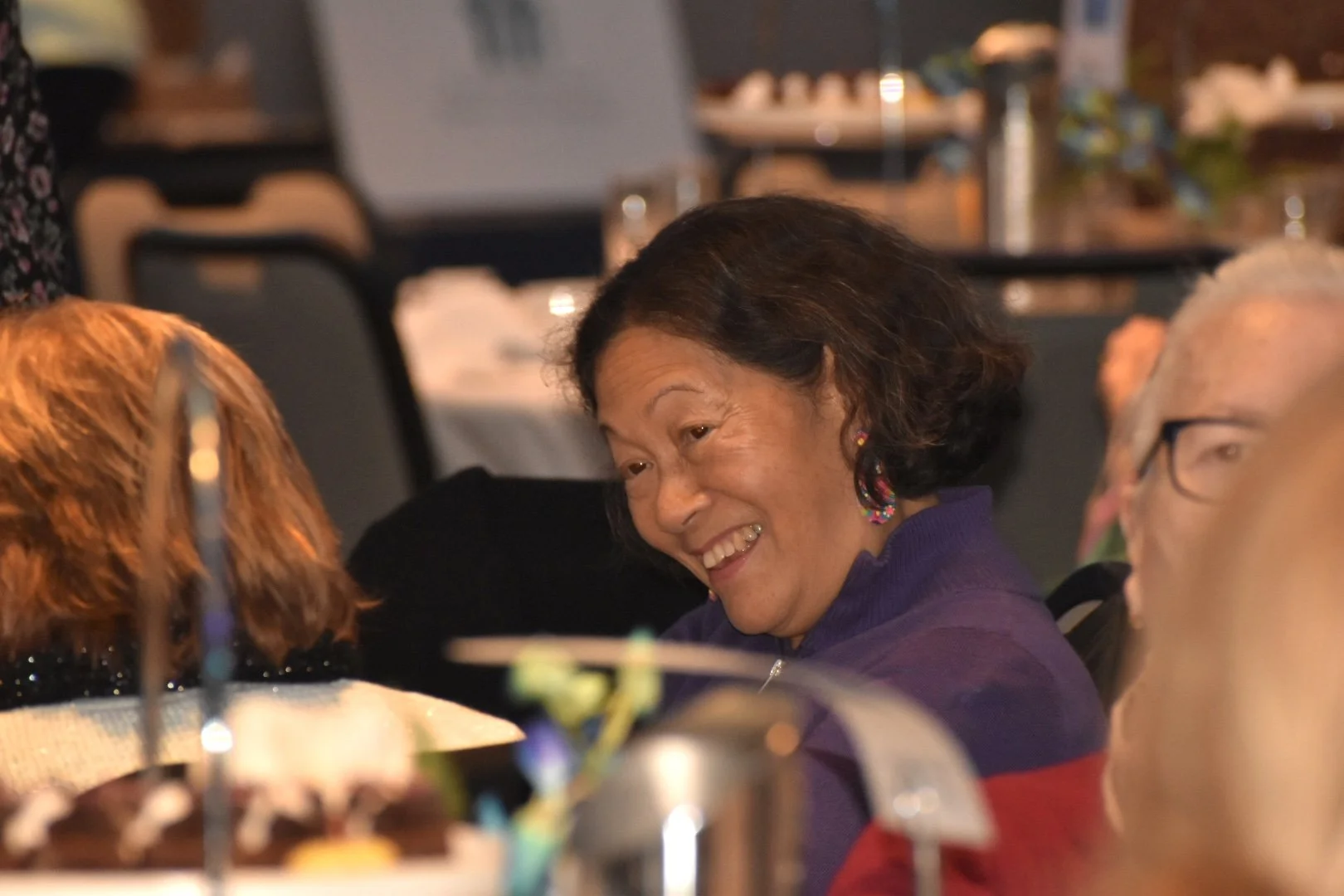 A woman laughing and talking with others at a social gathering or event, with a man visible on her right and other blurred figures and tables in the background.