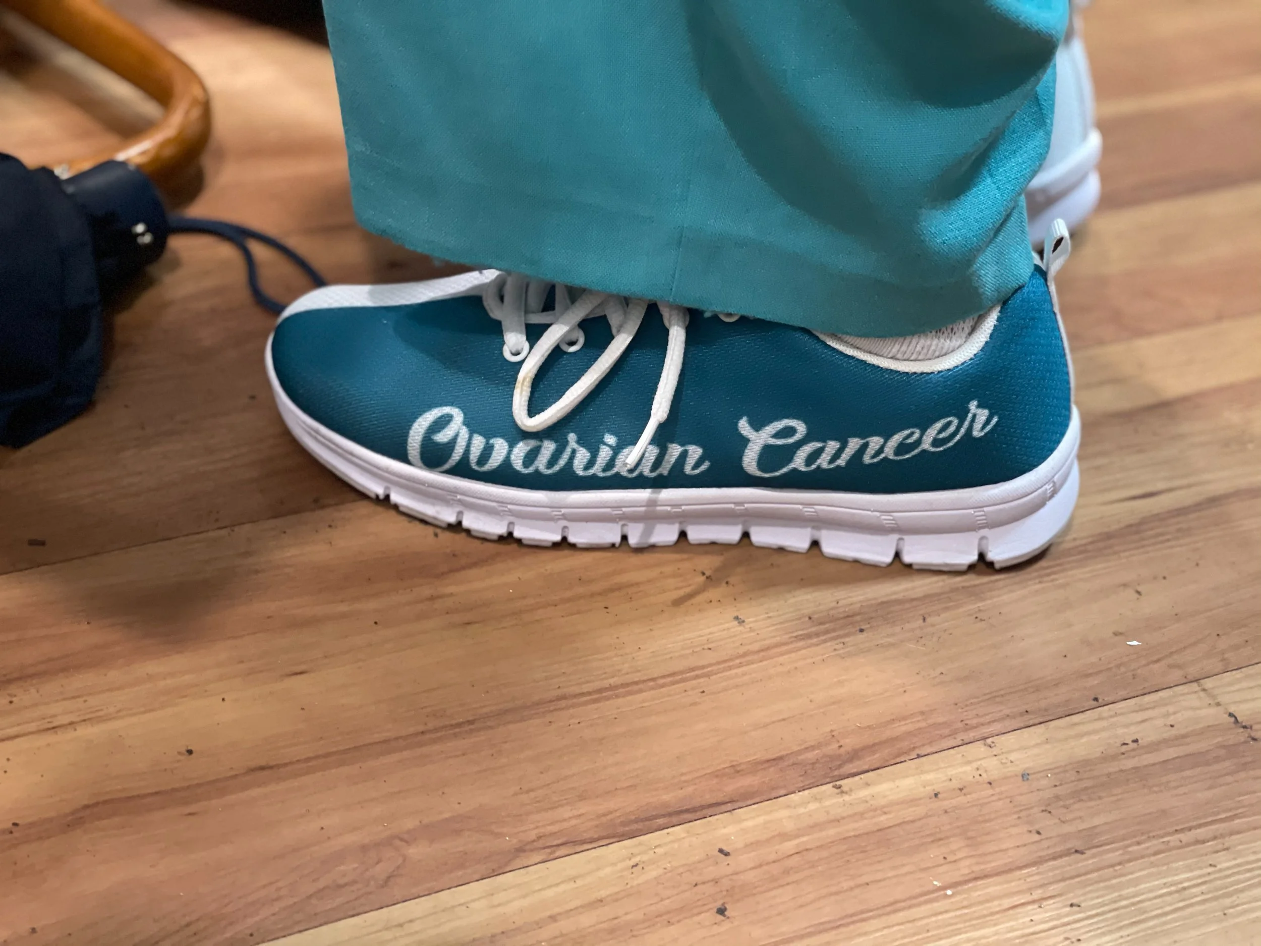 A teal sneaker with white laces and sole, with the words 'Ovarian Cancer' written on the side, worn by a person standing on a wooden floor.