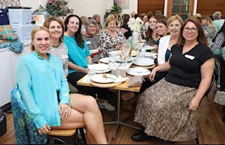 Group of women sitting around a table at a gathering or meeting in a room with decorated walls and a computer desk in the background.