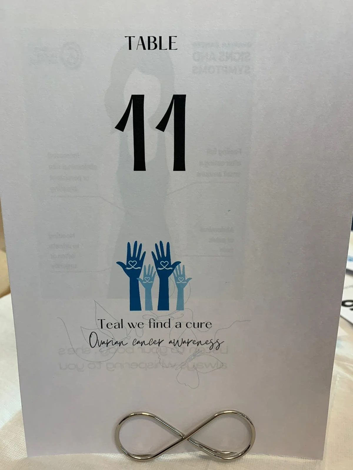 Table number 11 with a teal awareness message for ovarian cancer, featuring blue hands with hearts, and a metallic infinity symbol at the bottom.