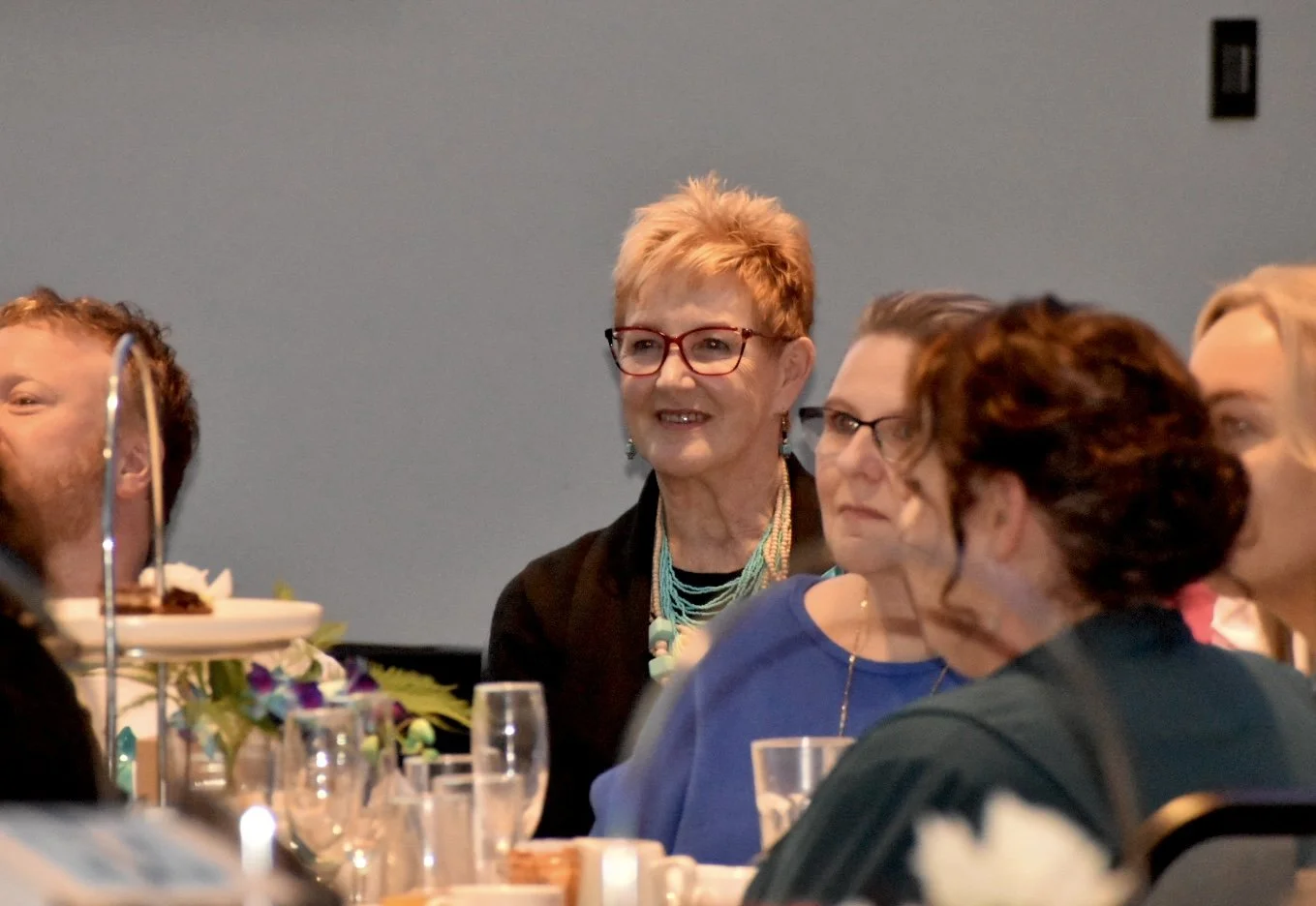 A group of people sitting at a banquet table during a formal event. The woman in the center has short, curly blond hair, red glasses, and is wearing a multi-strand turquoise necklace. She is smiling and looking ahead. Other guests are visible next to