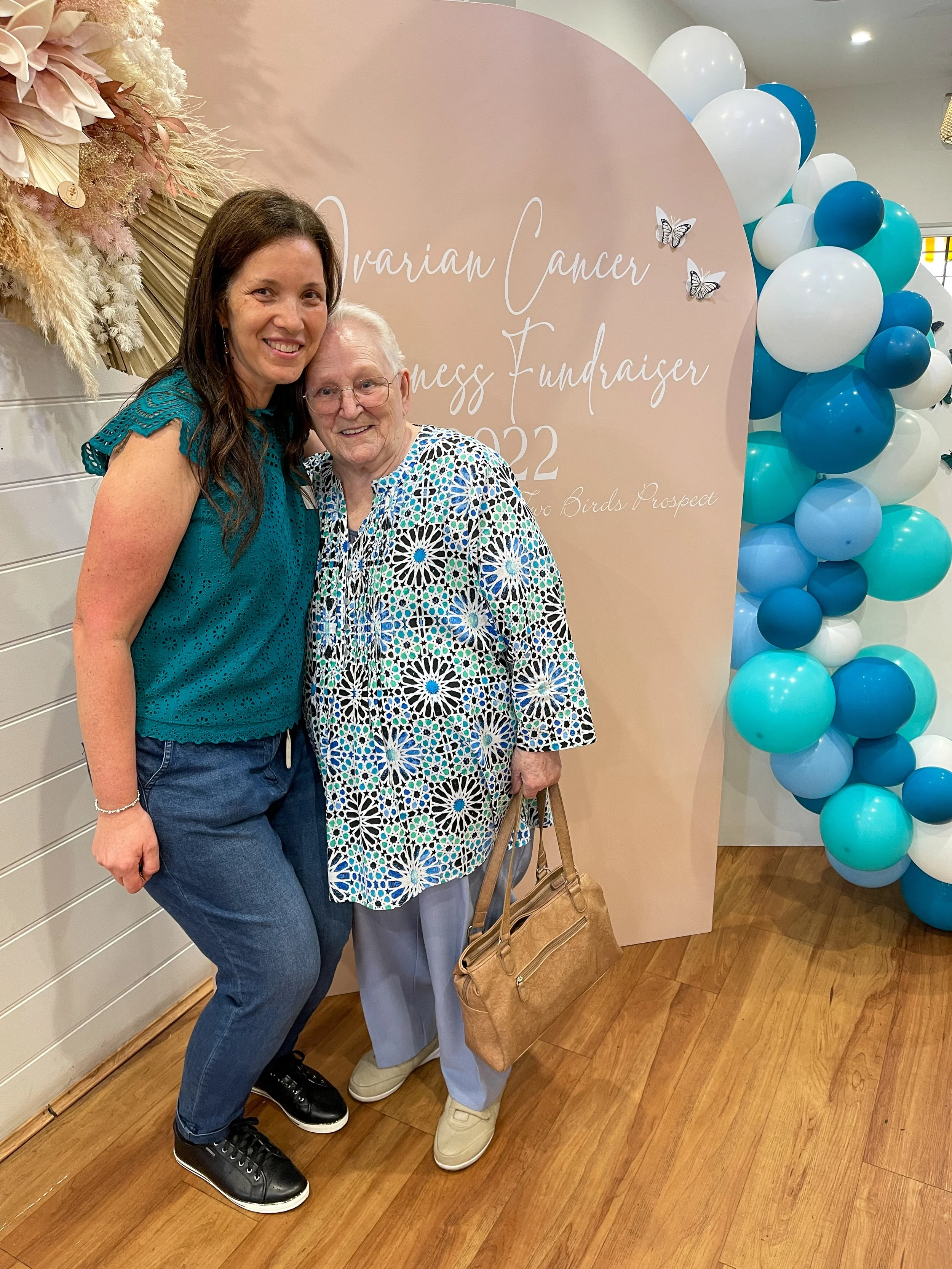 Two women standing together at a breast cancer awareness fundraiser, smiling. Behind them is a pink decorated backdrop with white and dark blue balloons on the side, and text that reads 'Ovarian Cancer Awareness Fundraiser 2022' and 'Love Birds Prosp