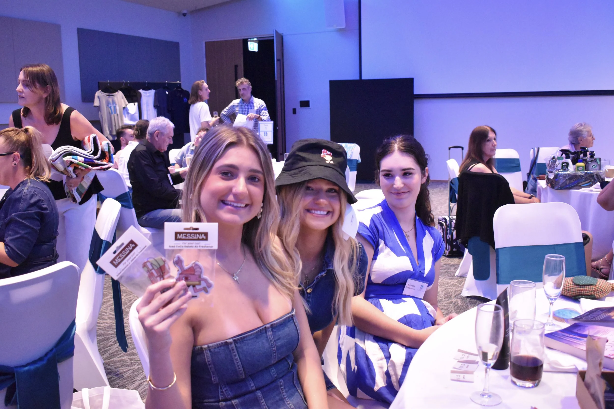 Three smiling young women sitting at a table during an event, with the woman on the left holding a pack of assorted souvenirs including a small horse figurine, and the background shows other attendees and event merchandise.