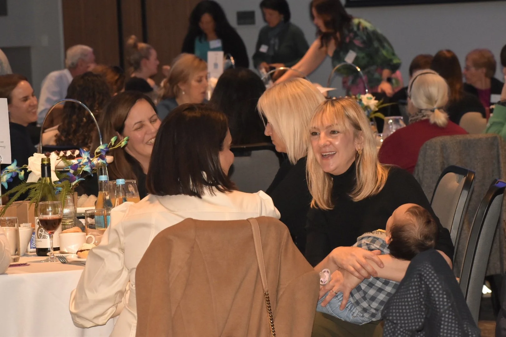 Women and children seated at round tables during a social event, engaged in conversation and smiling.