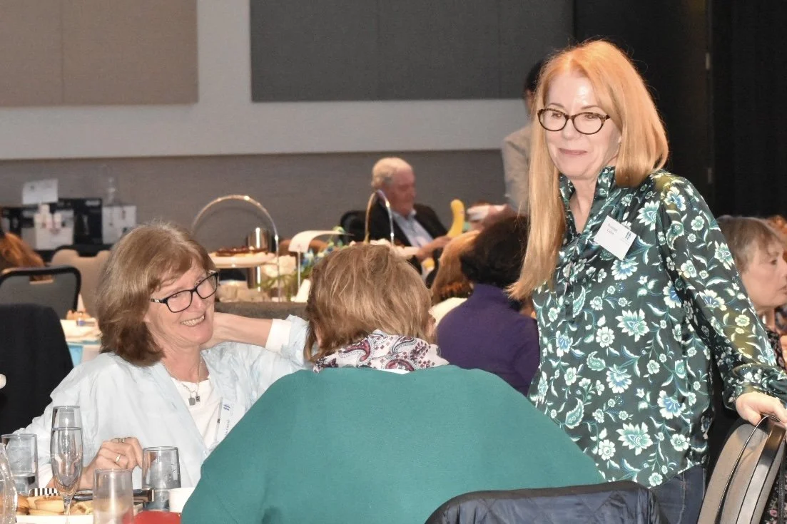 Women talking and smiling at a social event in a banquet hall, with tables, drinks, and other attendees in the background.