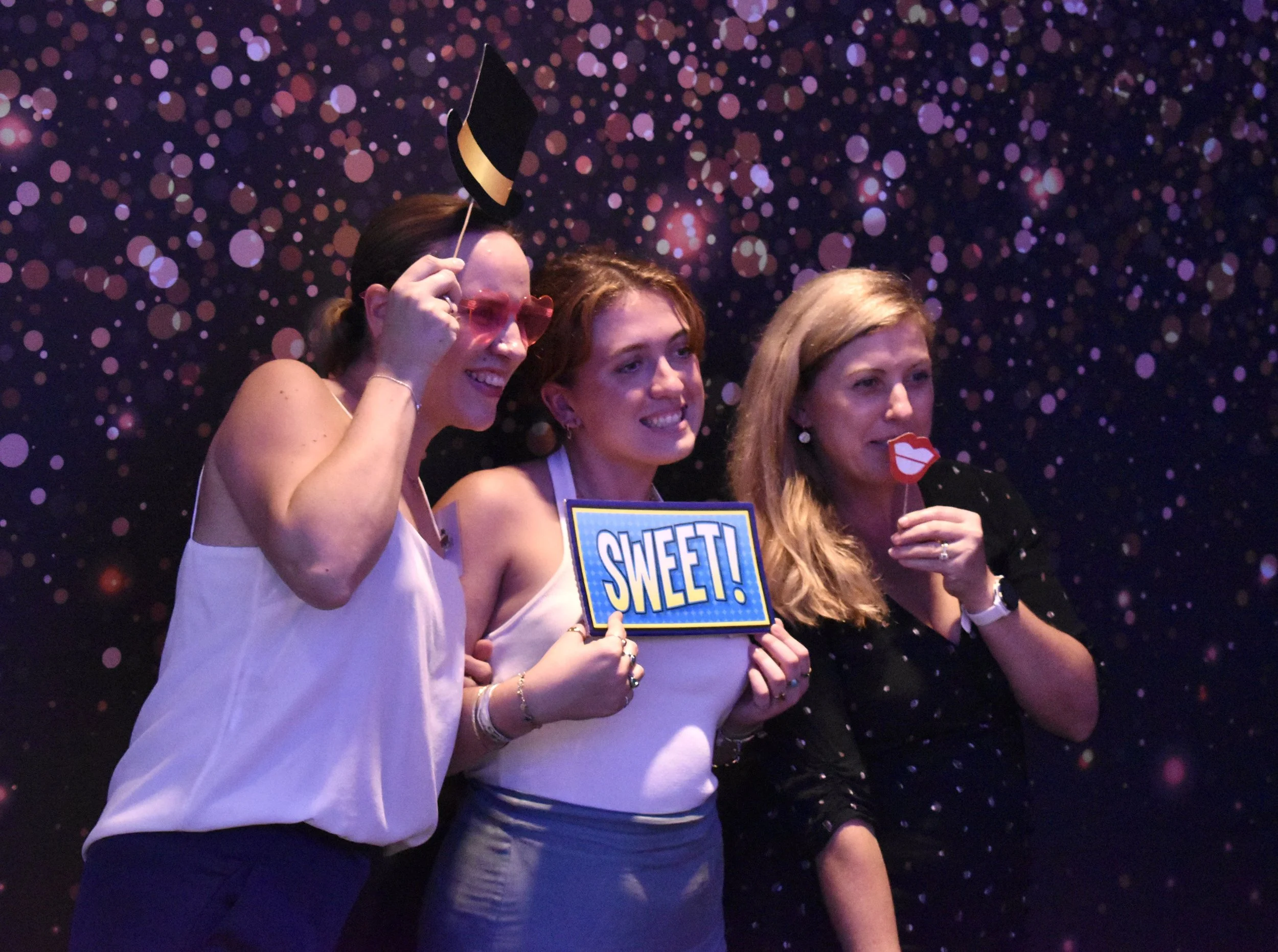 Three women at a photo booth having fun, holding playful props like a sign that says "SWEET!", a Lips prop, and a hat, with a colorful light display background.