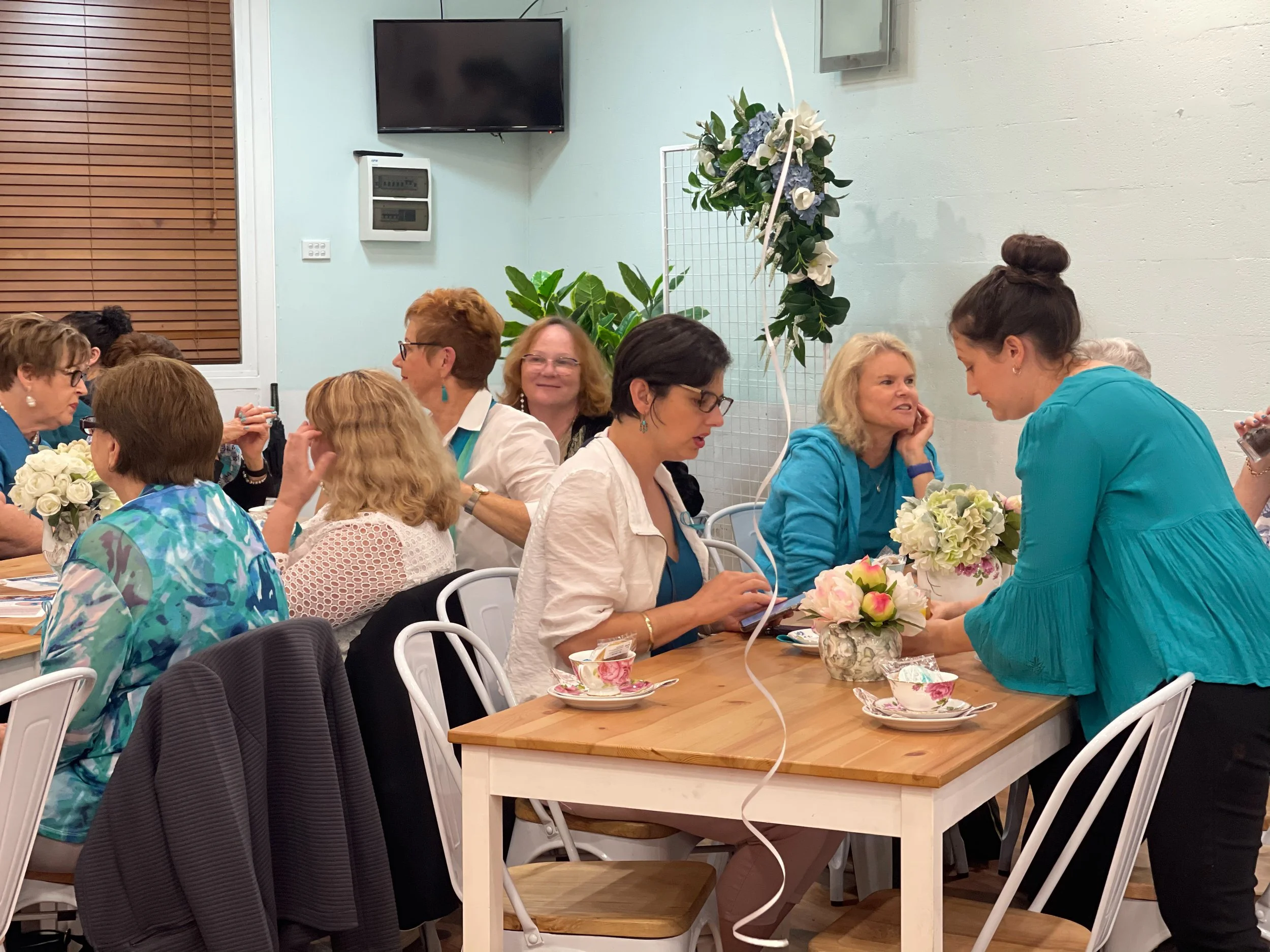 People sitting at a table decorated with flowers, engaged in conversation, in a brightly lit room with white walls, a wall-mounted TV, and a large window with wooden blinds.