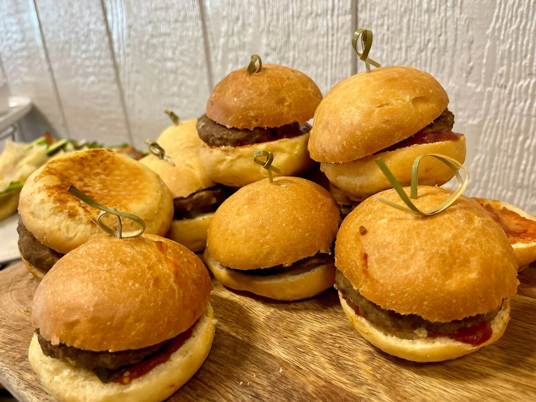 Mini hamburgers arranged on a wooden serving board, featuring buns with beef patties, some with ketchup and others with cheese, skewered with toothpicks.