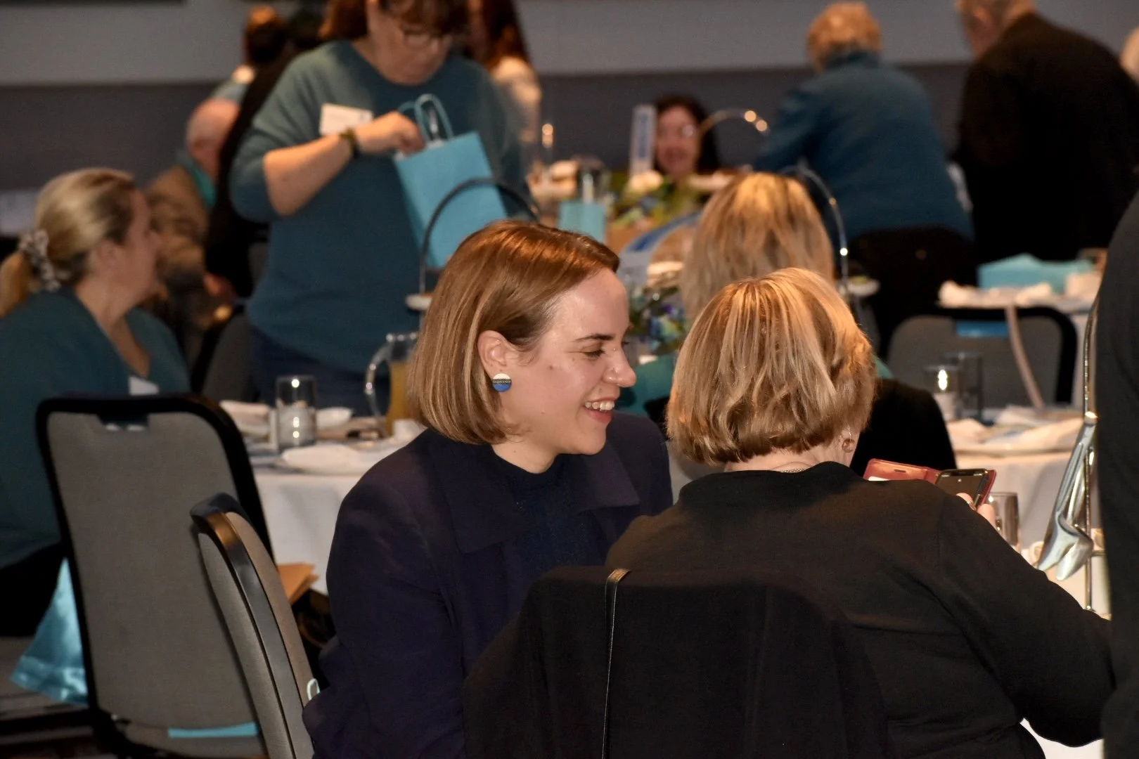 People sitting at round tables in a conference or banquet setting, engaging in conversation and examining their phones, with gift bags and glasses on the tables.