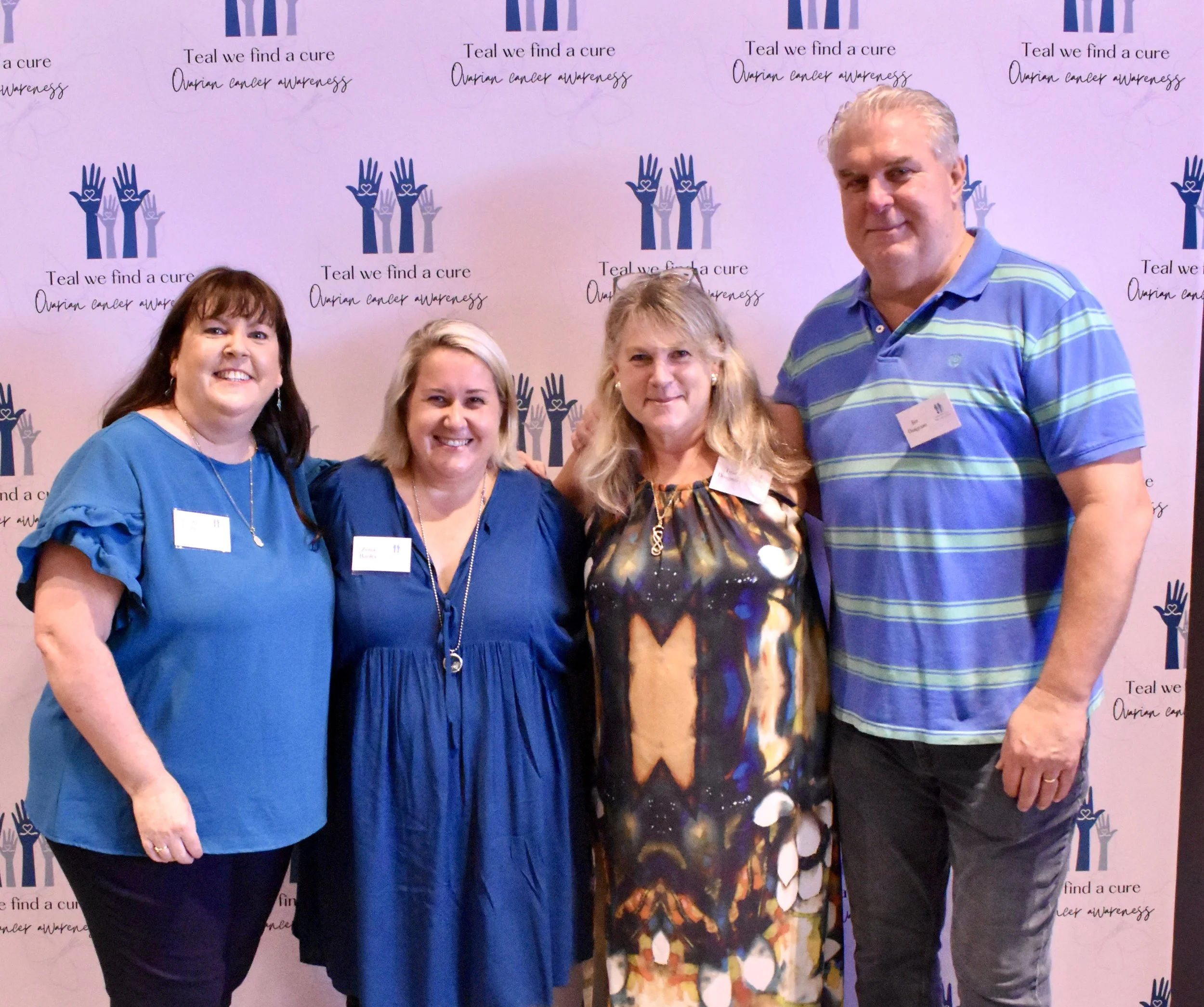 Four smiling women standing in front of a pink backdrop with blue and purple graphics and the text 'Teal we find a cure, Ovarian cancer awareness.'