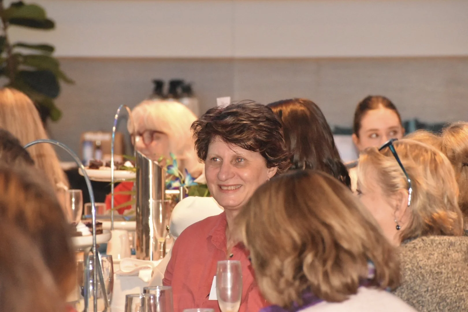 A group of women seated at a dinner table, smiling and engaged in conversation. The table is set with drinks, plates, and decorative elements, suggesting a social or celebratory event.