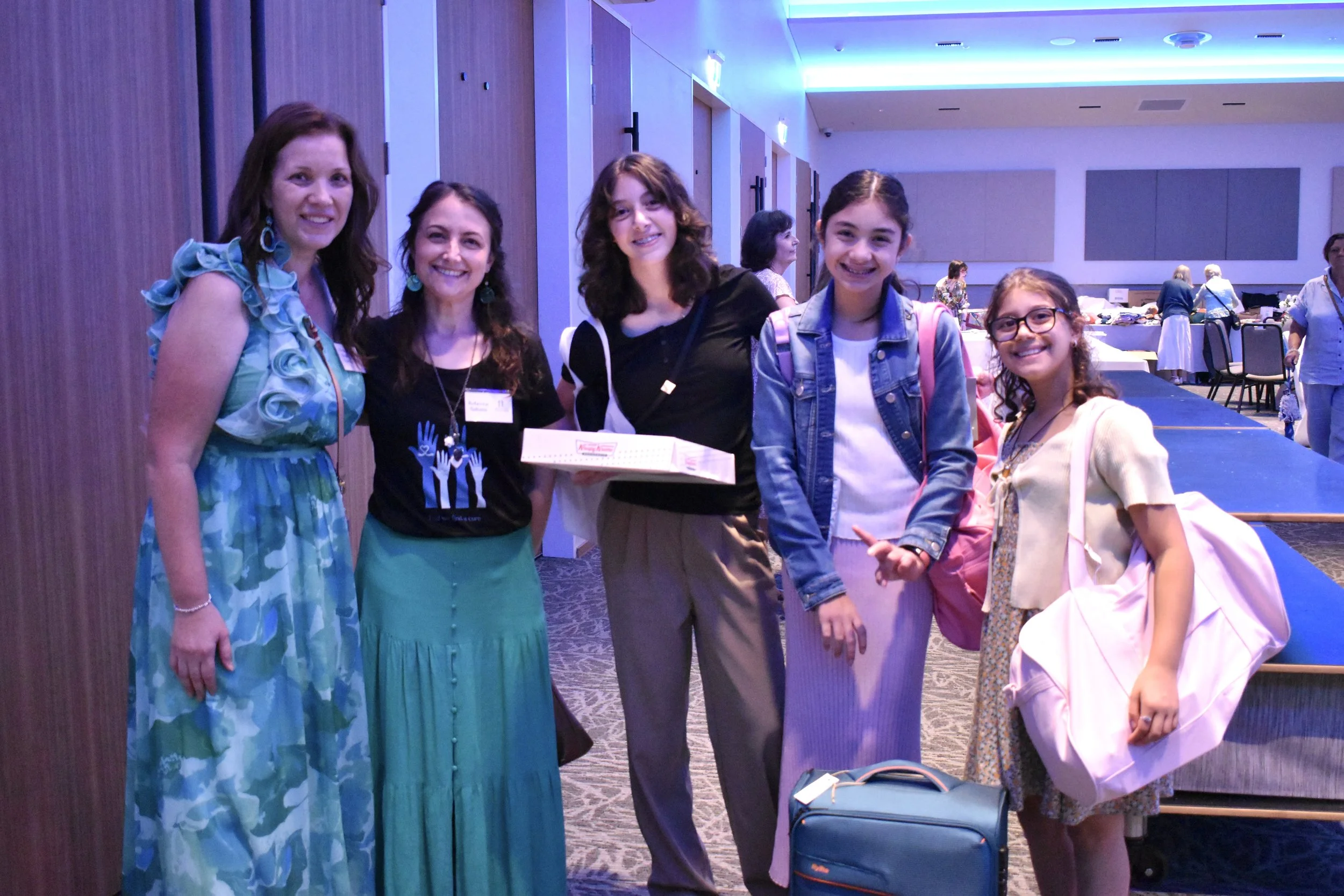 Group of five women and girls standing in a conference room, smiling at the camera, with tables and other people in the background.