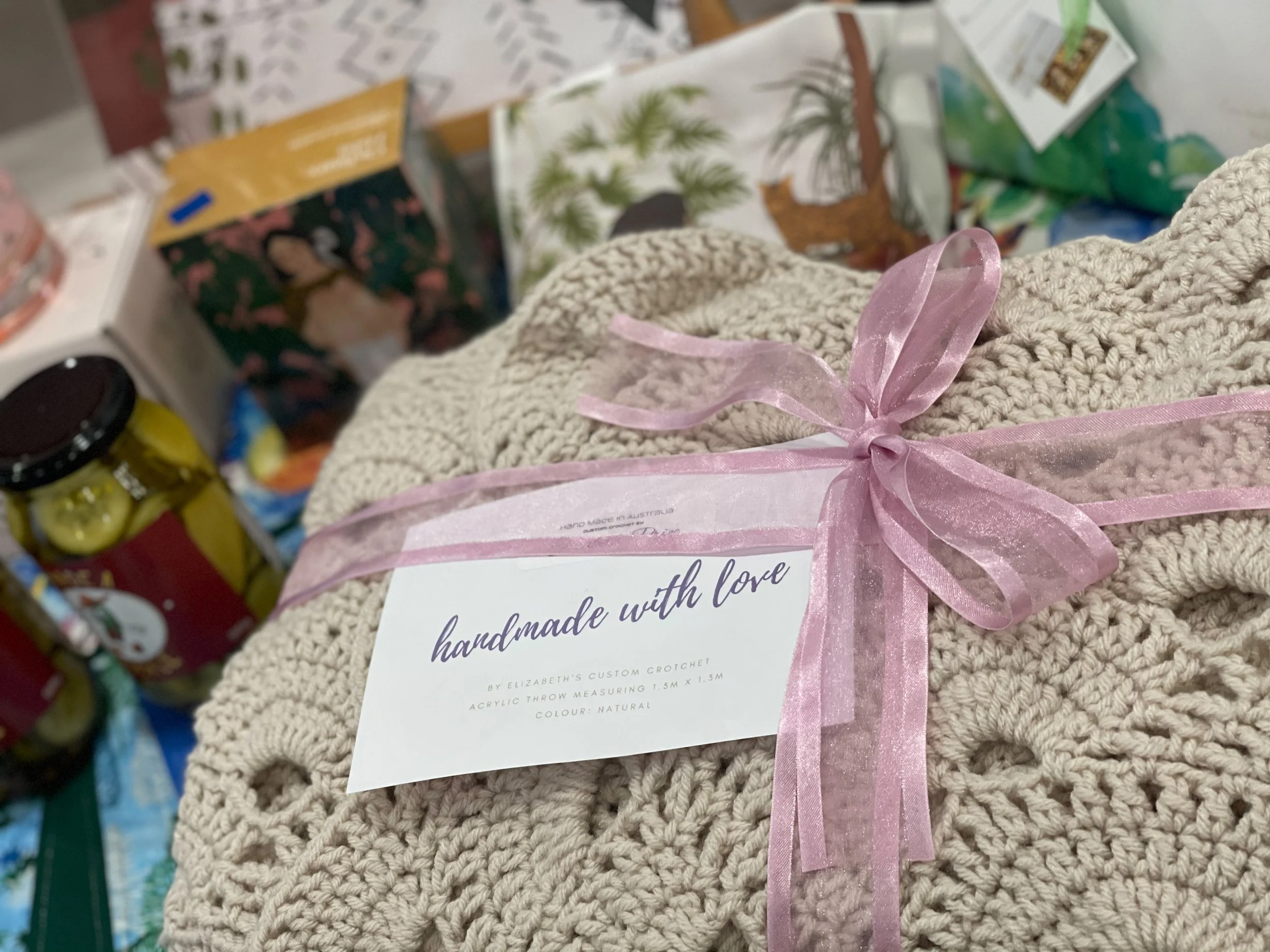 A beige knitted blanket with a pink ribbon bow, labeled 'handmade with love' and indicating it is an acrylic throw measuring 1.5 meters by 1.3 meters, displayed among various food and gift items.