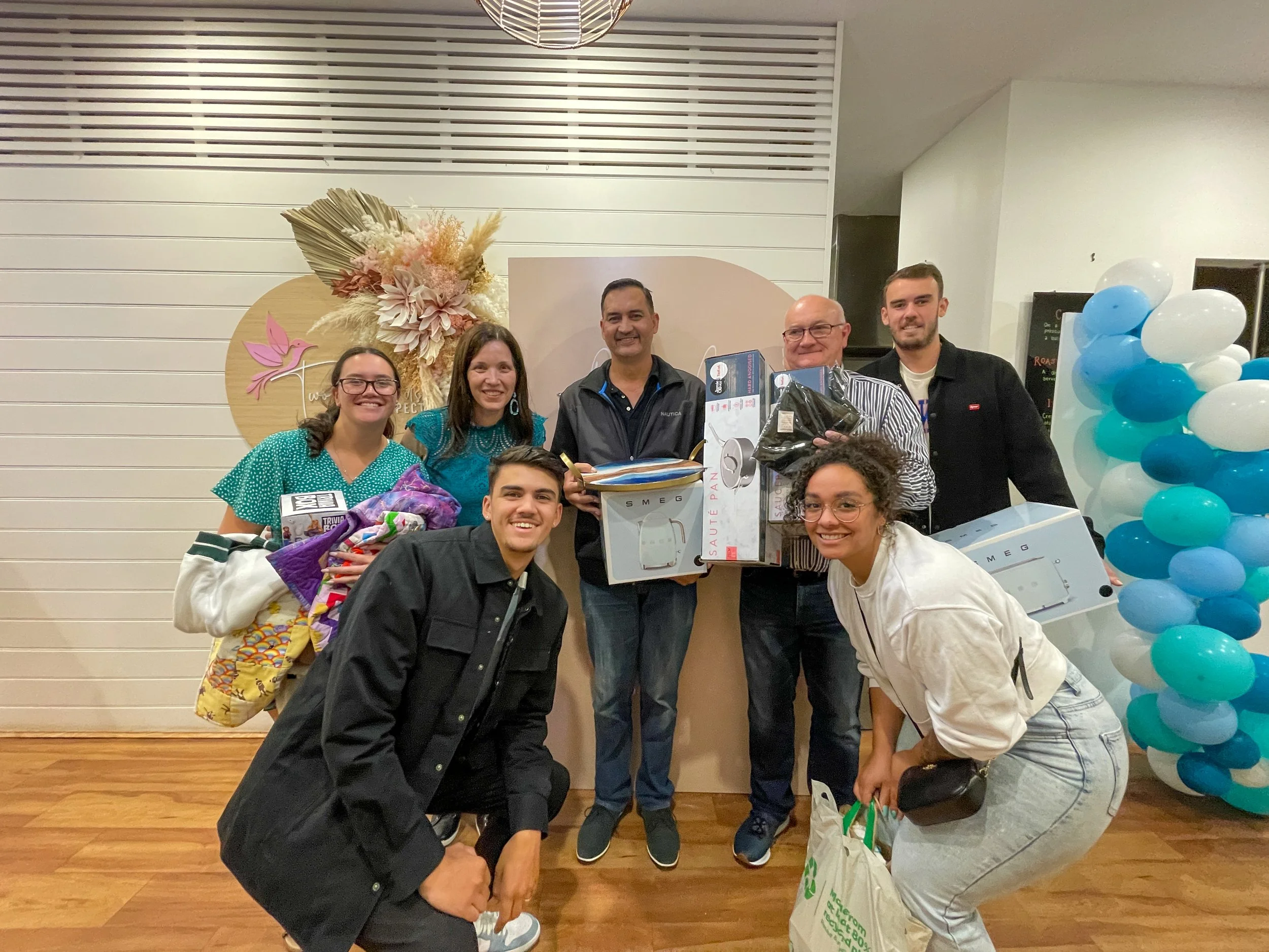 A group of nine people posing together at an indoor event, holding gifts and smiling, with a decorative floral wall and balloons in the background.