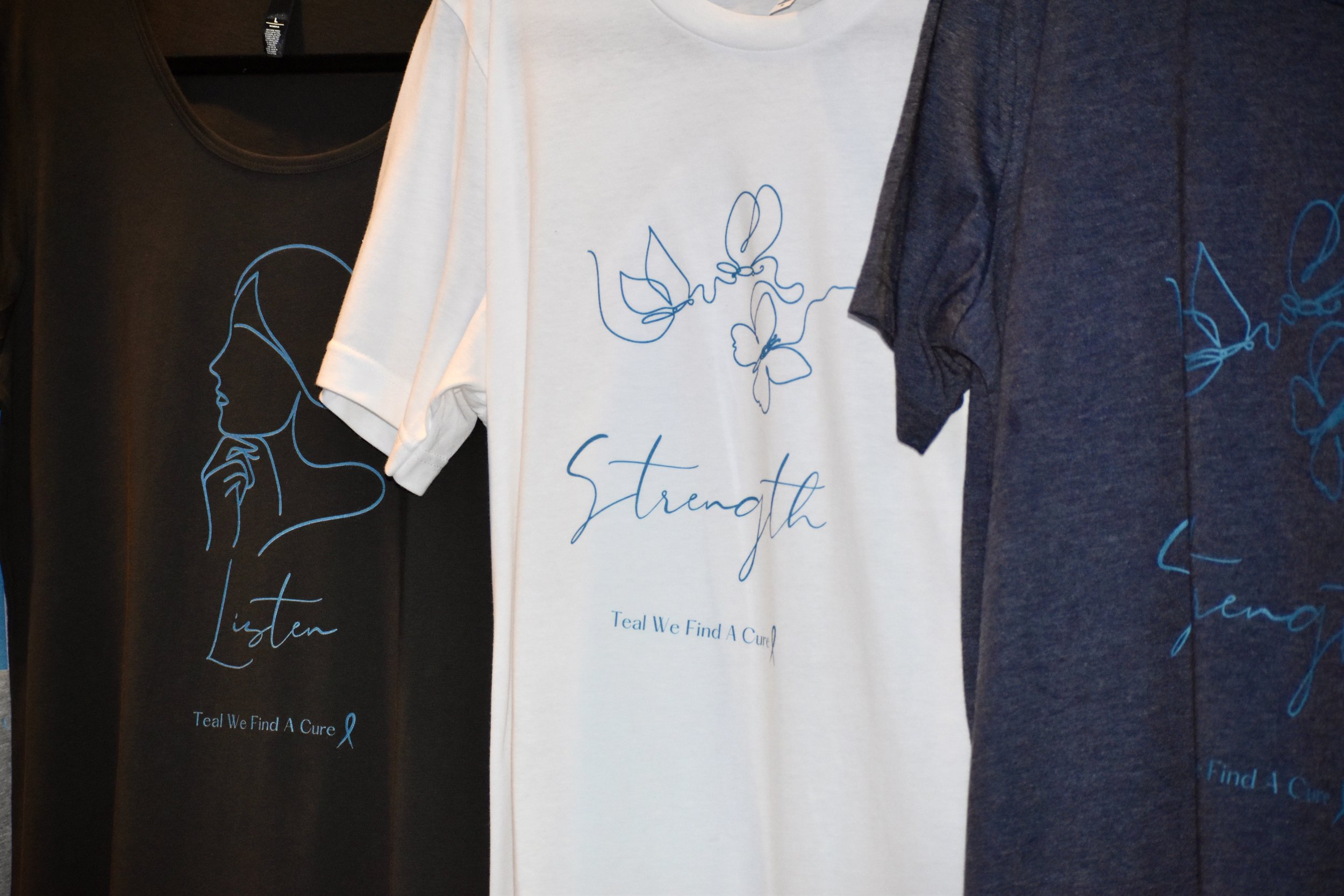 White and dark gray T-shirts hanging on display, featuring butterfly and woman's profile line art designs with words 'Strength' and 'Listen', and the phrase 'Teal We Find A Cure' with a teal ribbon symbol.