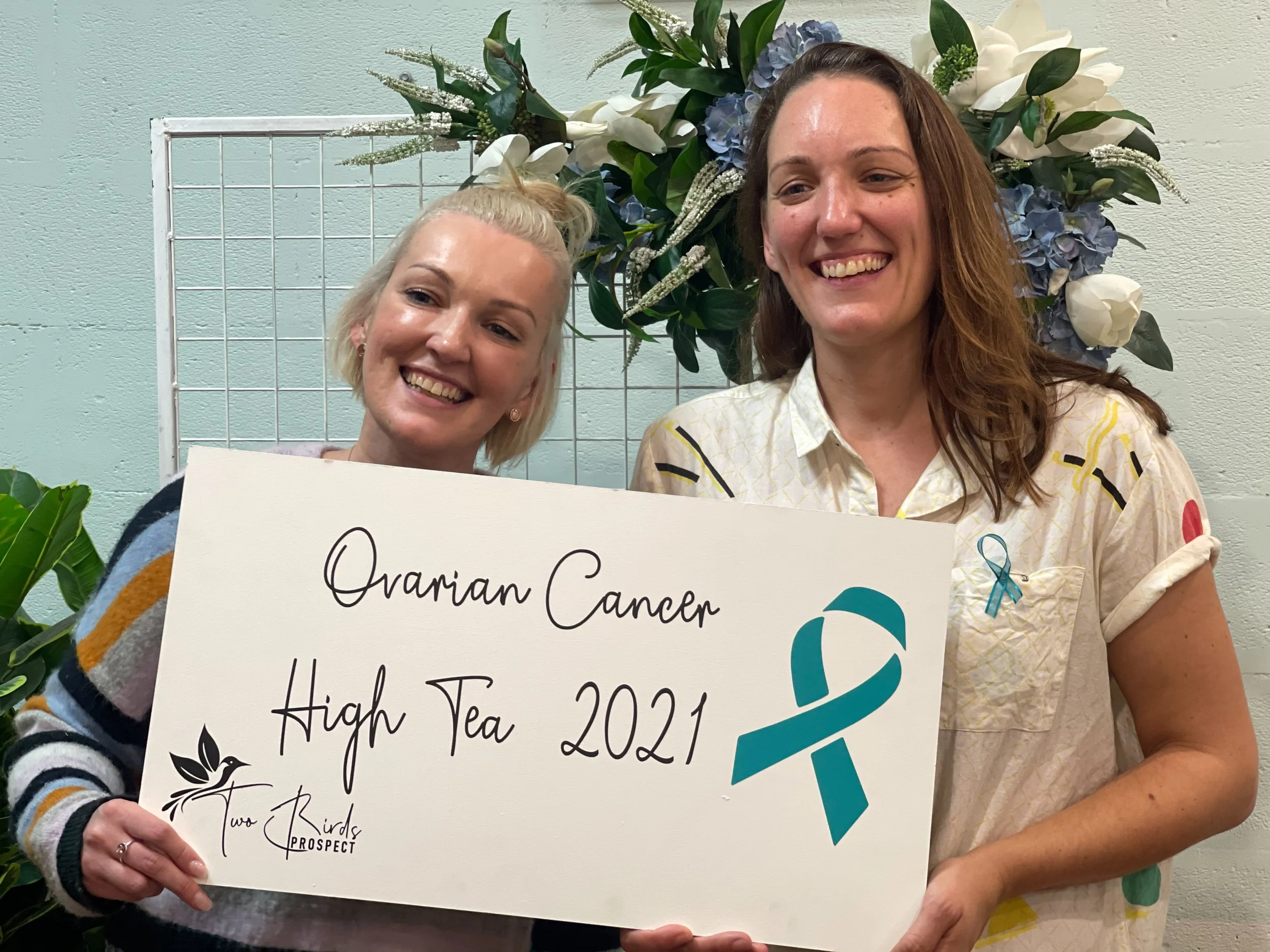 Two women smiling, holding a sign that reads 'Ovarian Cancer High Tea 2021' with a teal ribbon and a logo that says 'Two Birds Prospect', standing in front of a floral arrangement with white, blue, and green flowers.