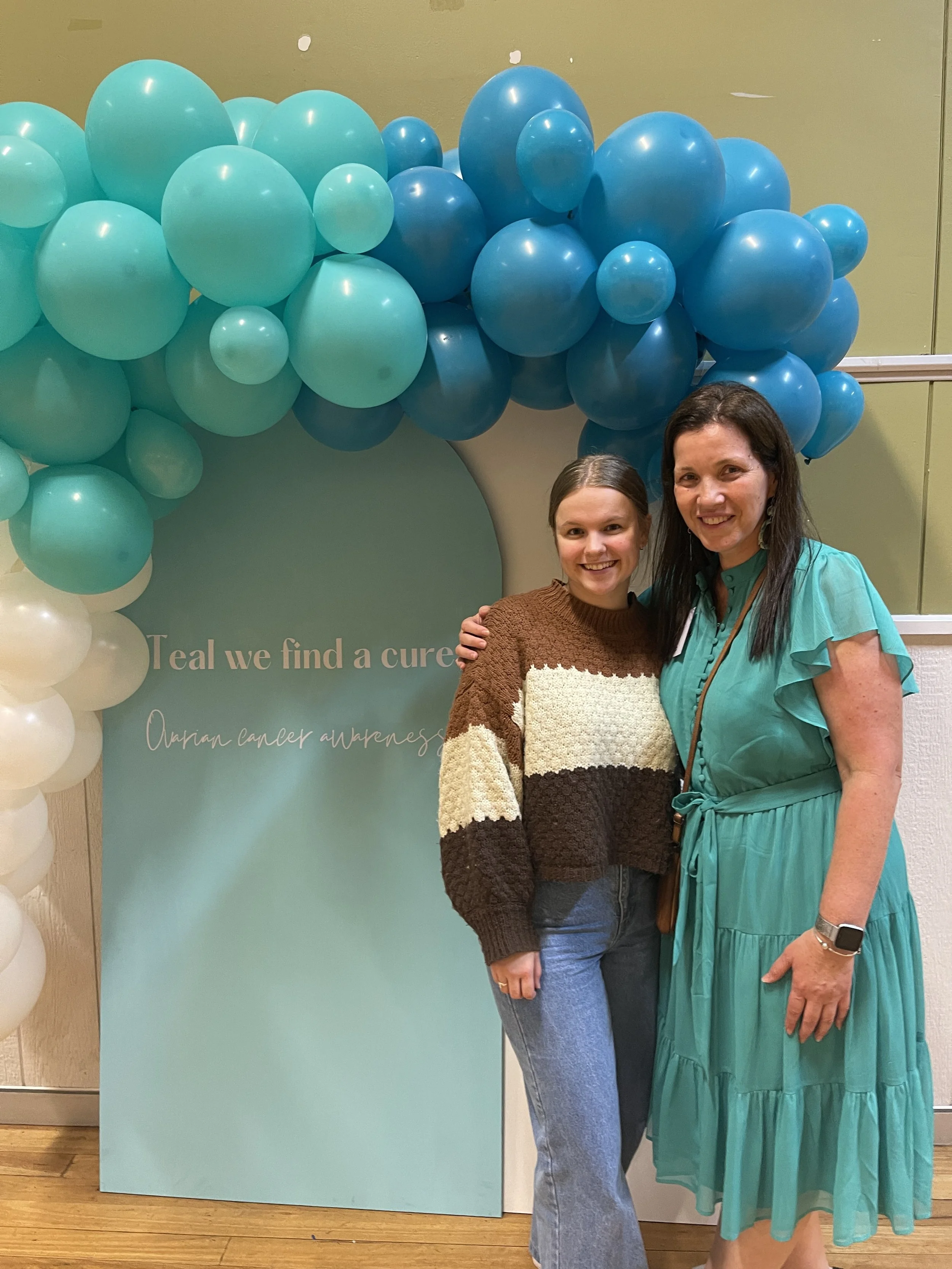 Two women smiling and posing in front of a teal balloon arch and a teal sign that reads 'Teal we find a cure' at a cancer awareness event.
