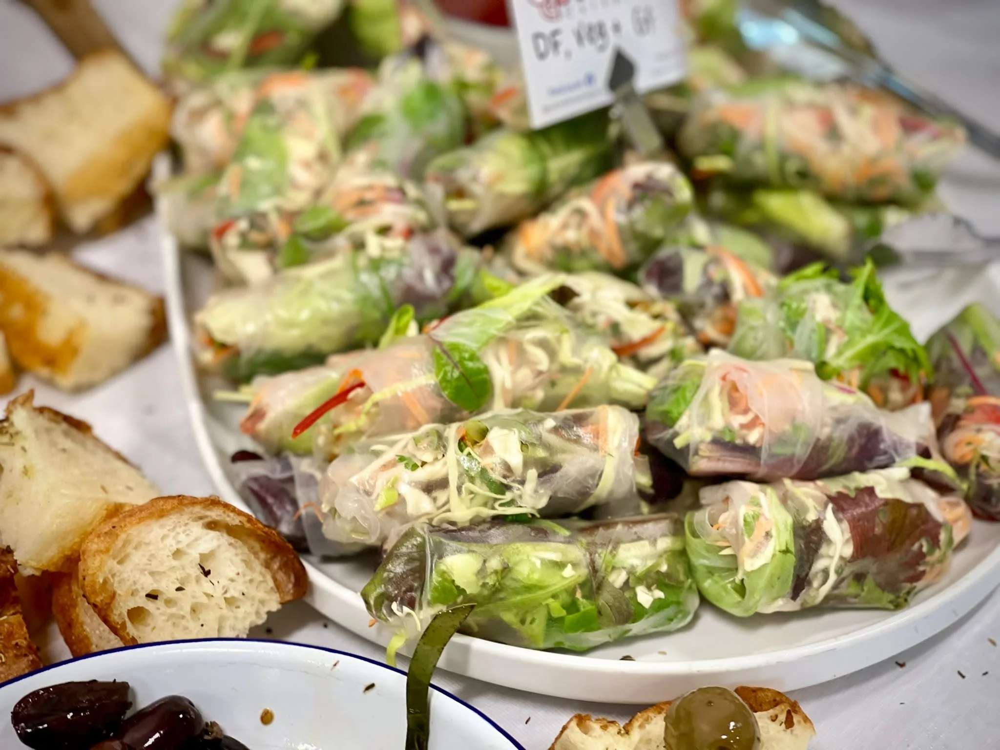 Close-up of a large white platter filled with fresh spring rolls wrapped in translucent rice paper, showing greens, vegetables, and possibly shrimp inside. Surrounding the platter are pieces of toasted bread and a small dish of black olives.