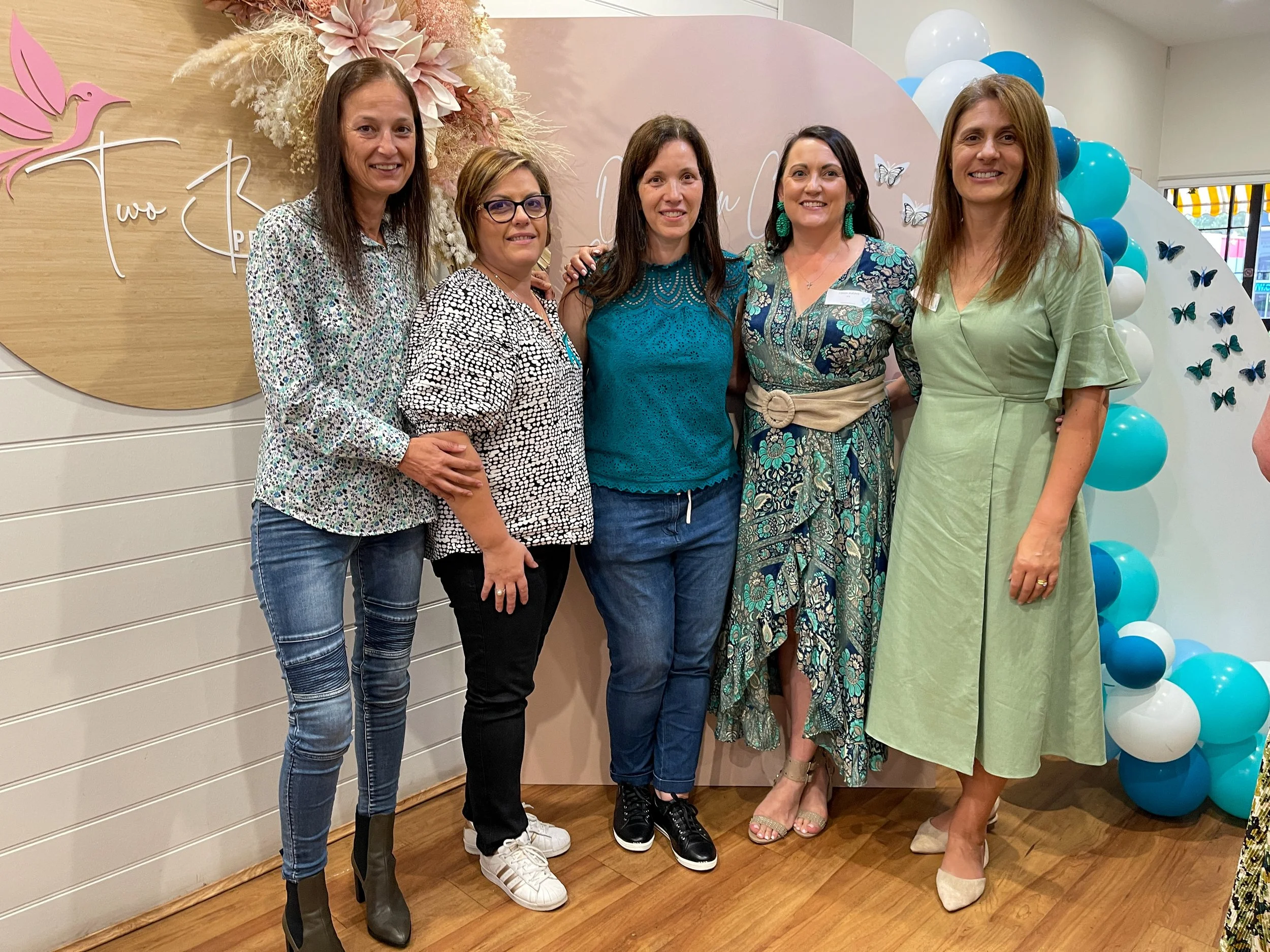 Five women standing together, smiling at an indoor event with pink and blue decorations, including a floral arrangement, butterflies, and balloons.