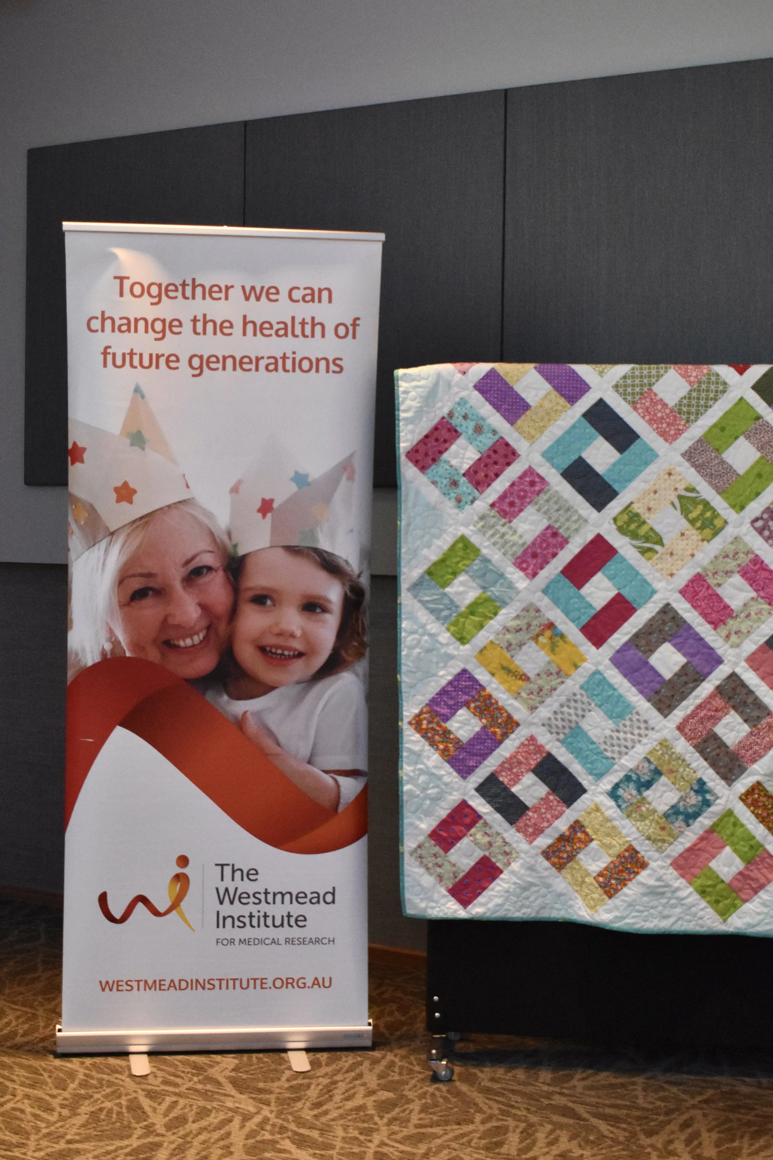 A banner standing next to a colorful quilt on display. The banner shows an image of a smiling woman and child wearing paper crowns and has the text "Together we can change the health of future generations" along with the logo and website for The West