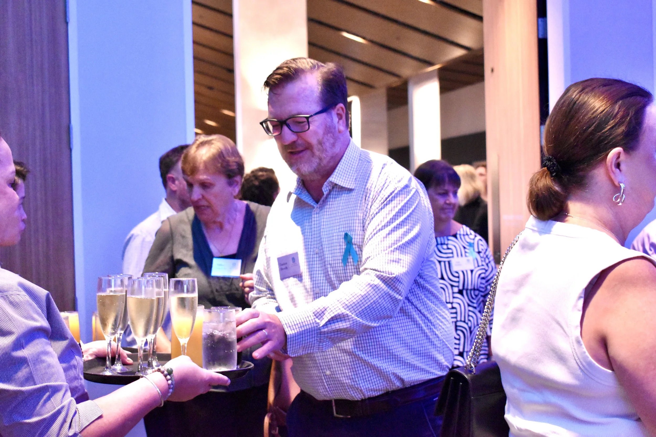 A man with glasses and a blue ribbon on his shirt receives a drink from a server at a social event, with several women in the background and a tray of champagne glasses in the foreground.