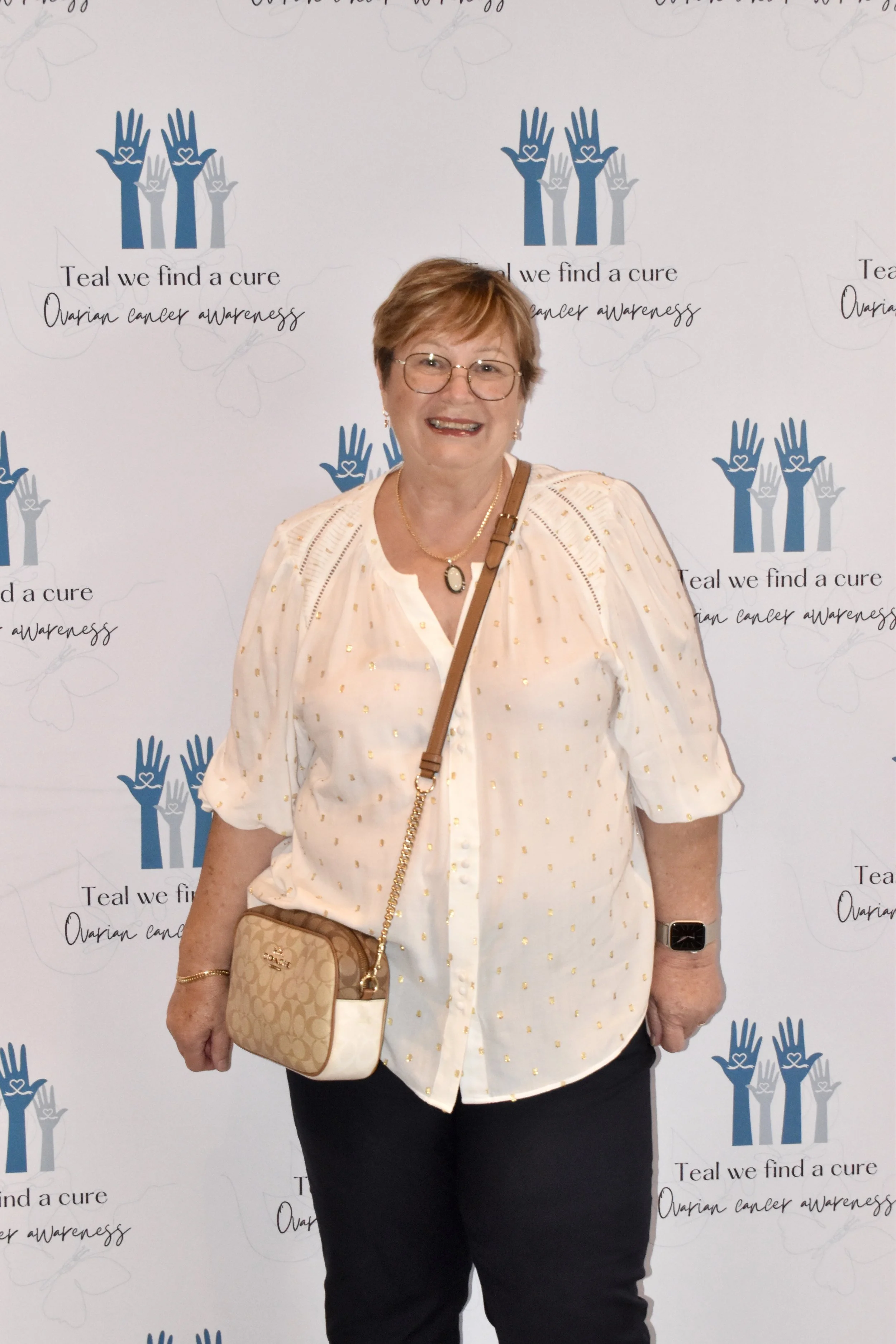 A woman with short hair and glasses smiling, wearing a cream blouse with gold polka dots, black pants, and carrying a beige Coach purse, standing in front of a step and repeat backdrop with blue hands and the text 'Teal we find a cure - Ovarian cance