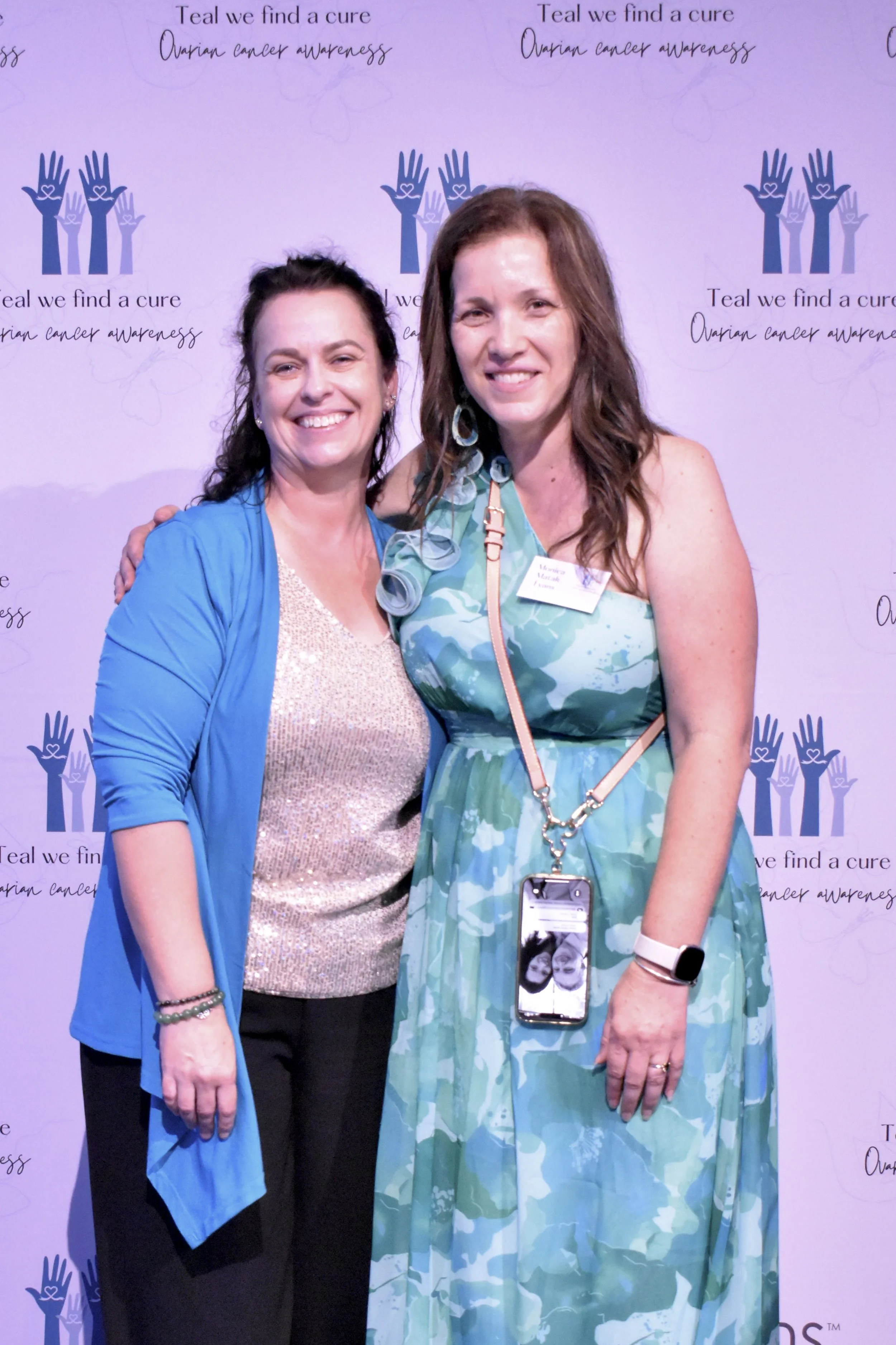Two women smiling and posing for a photo at an event with a purple backdrop that has the text "Teal we find a cure ovarian cancer awareness" and a logo of multiple hands with hearts.
