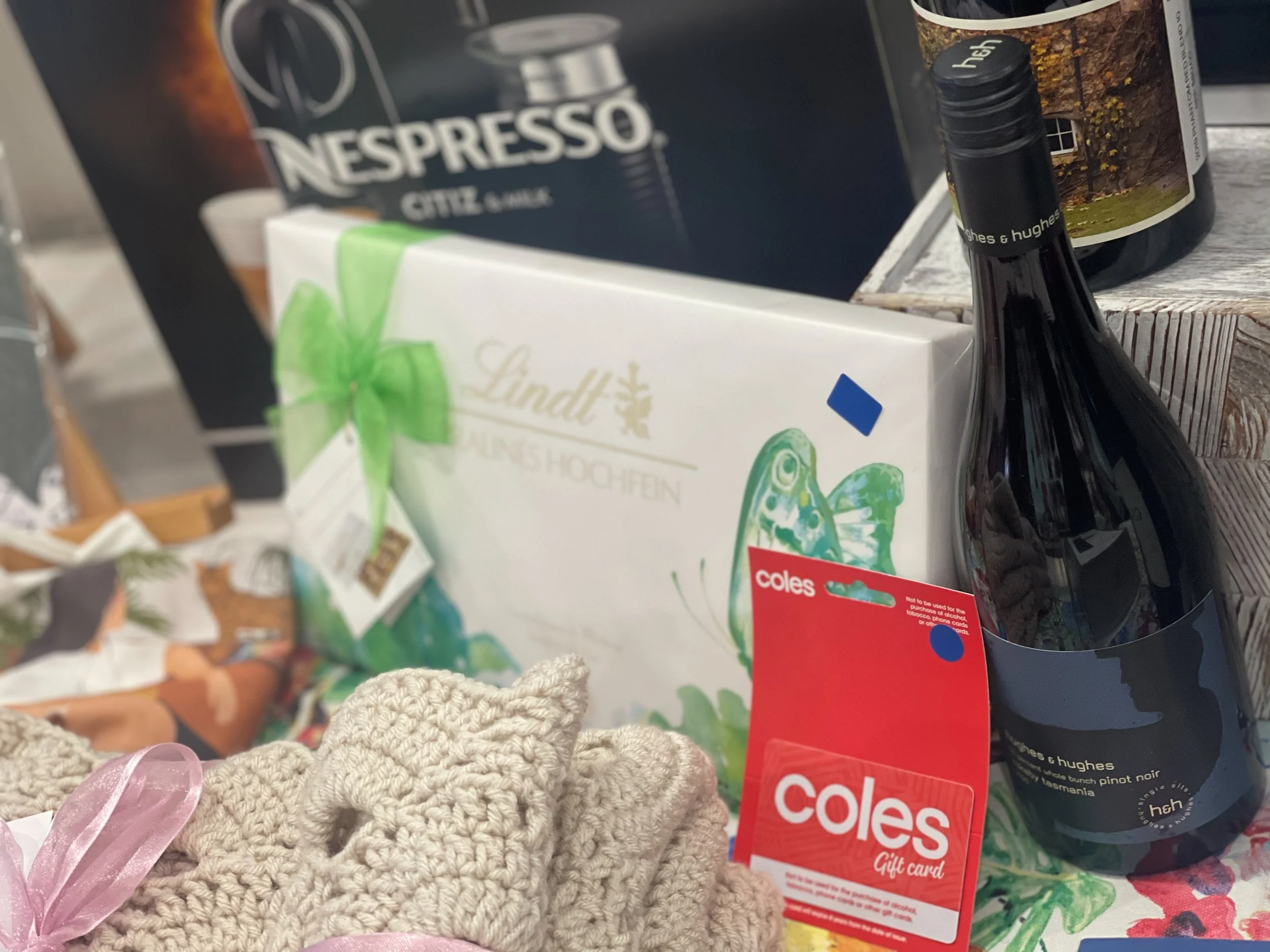 A gift basket containing Lindt chocolate, a bottle of wine, and a Coles gift card, with party favors including knit fabric items with pink ribbons in the foreground.