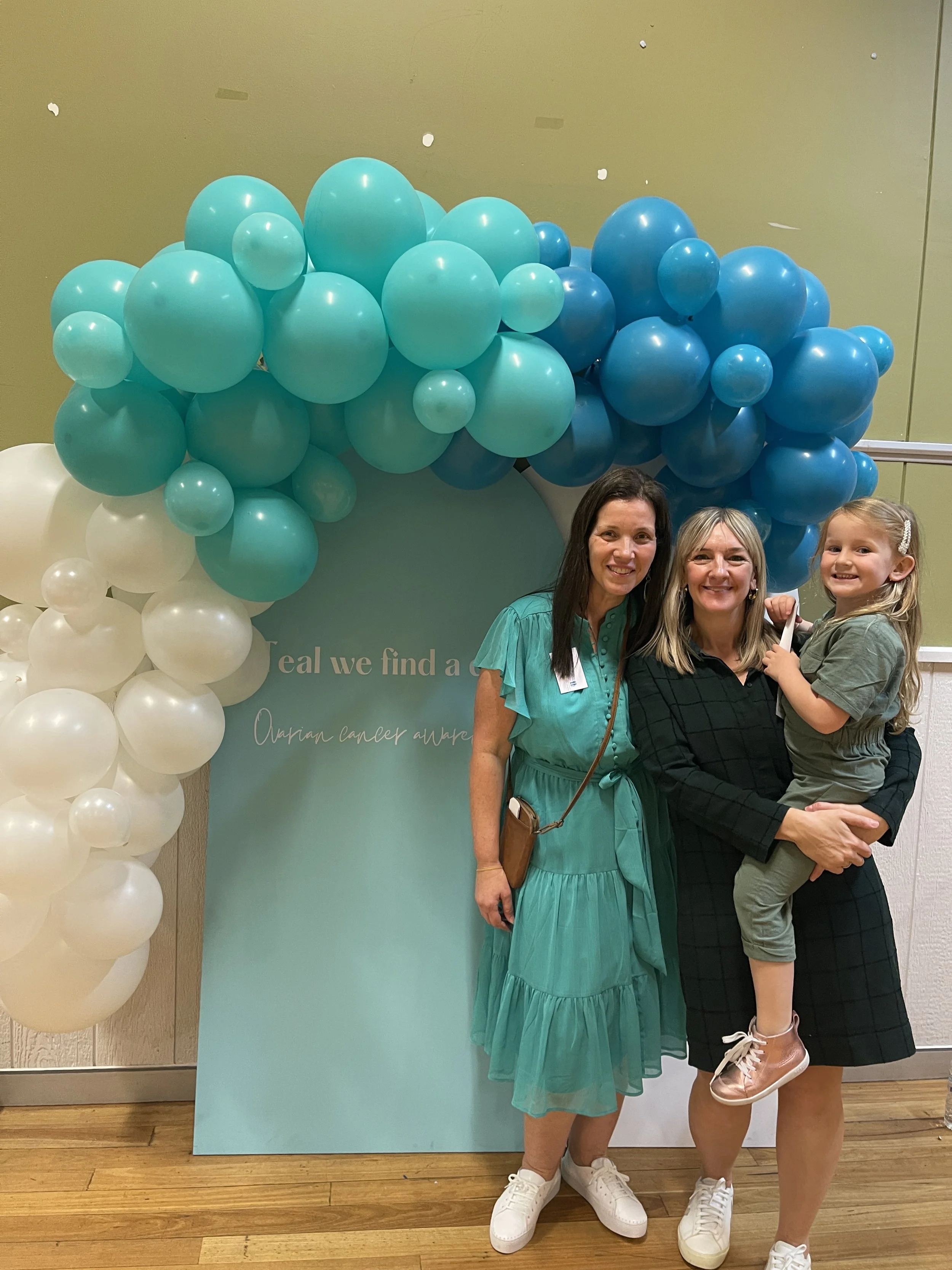 Three women and a girl standing in front of a teal-colored display with white and teal balloons, smiling at the camera.