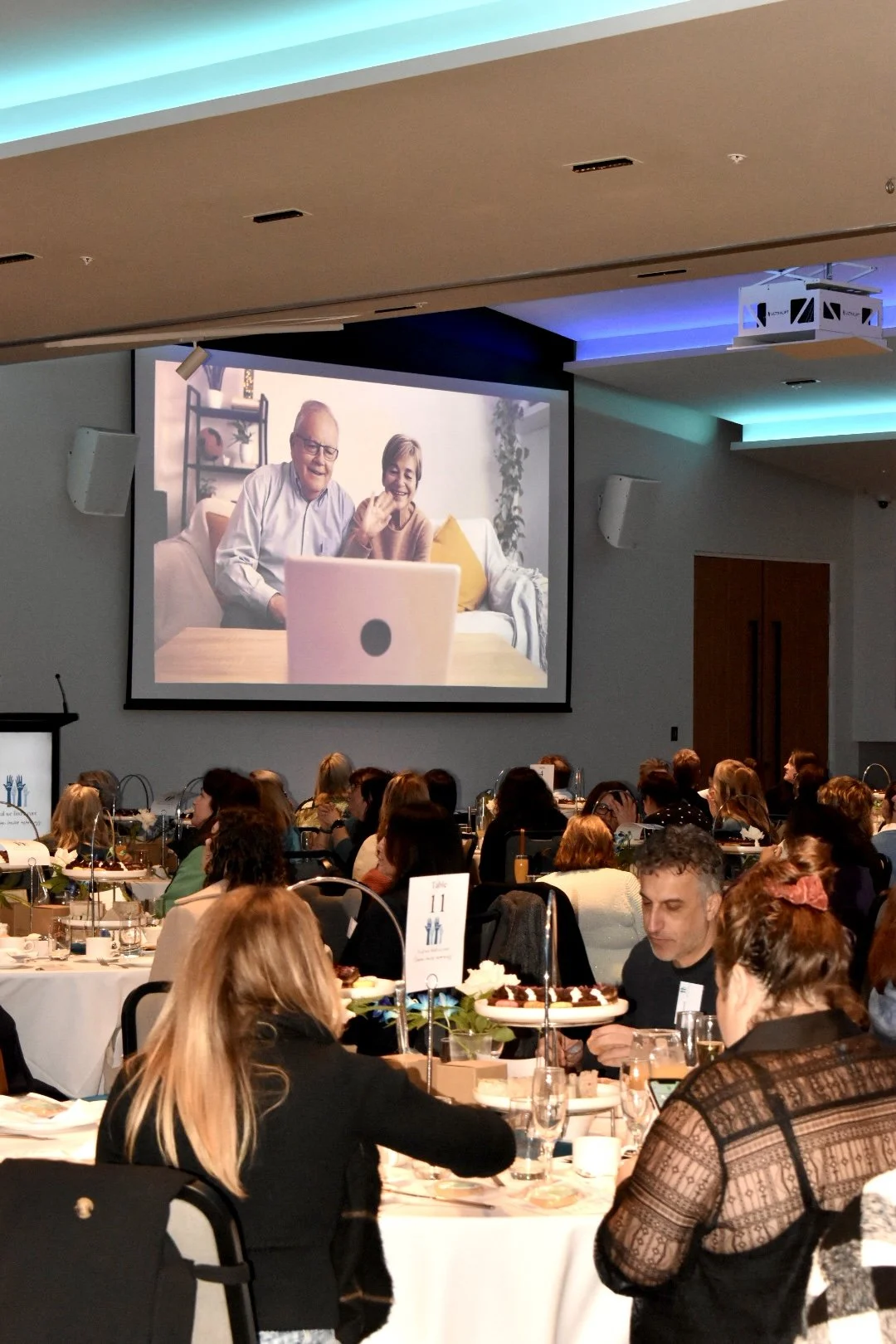 People attending a large conference or banquet in a hotel or conference center, watching a large screen displaying a video call with an elderly couple waving.
