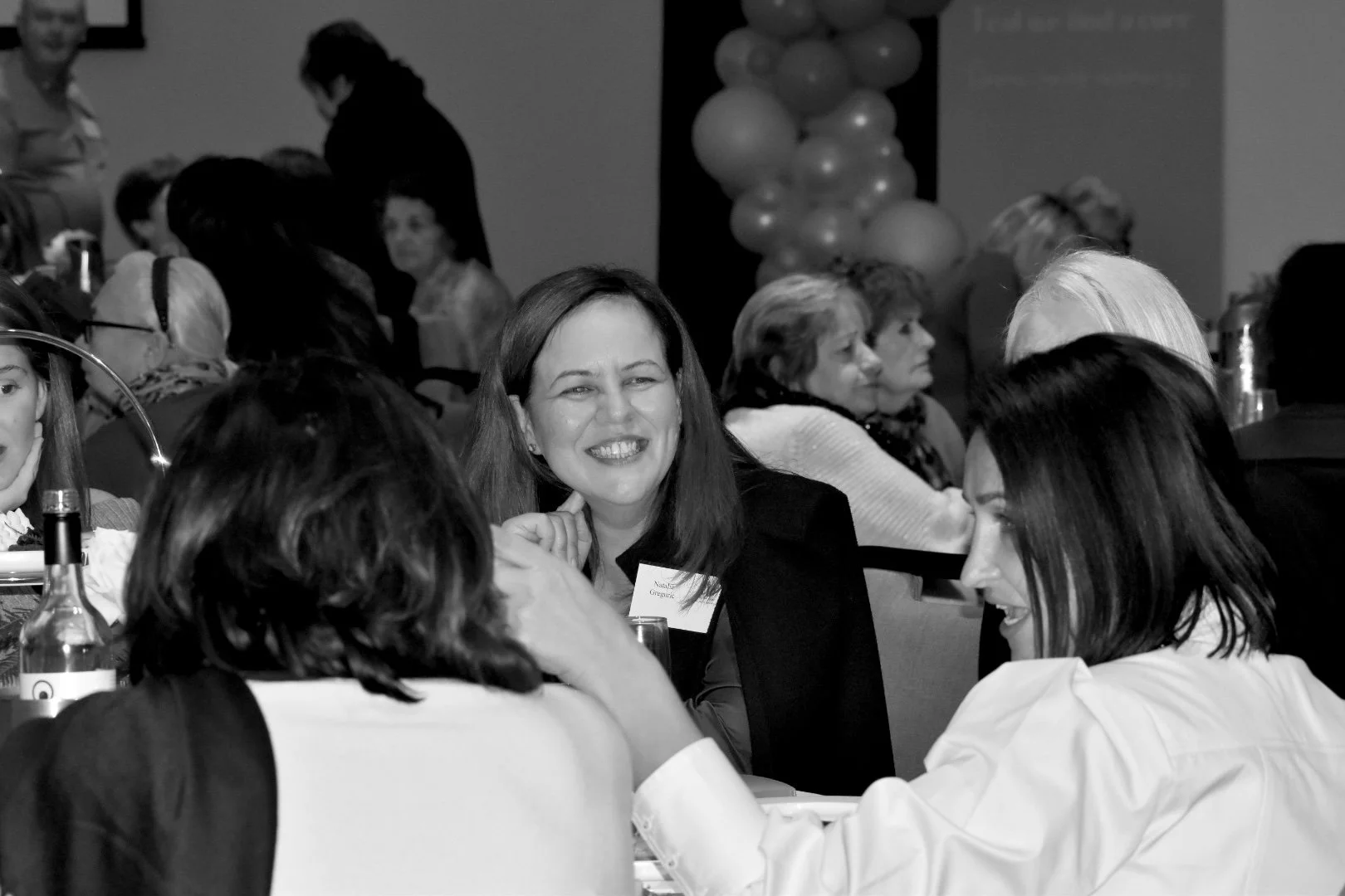A group of women sitting at a round table, engaged in conversation during an event. One woman in the center is smiling at the camera. The setting appears to be a formal or professional gathering.