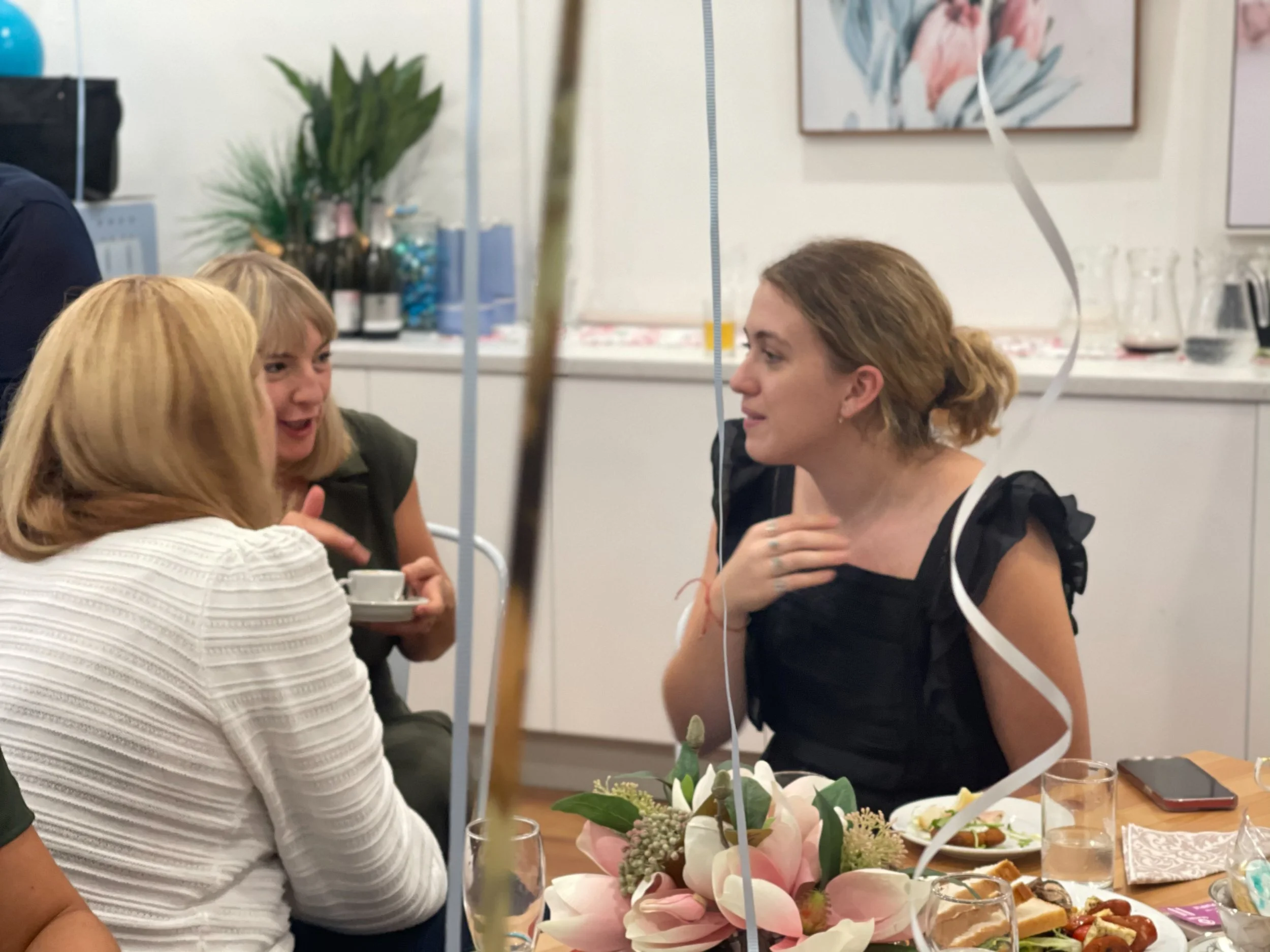 Group of women engaging in conversation at a decorated party table with flowers, food, and drinks.