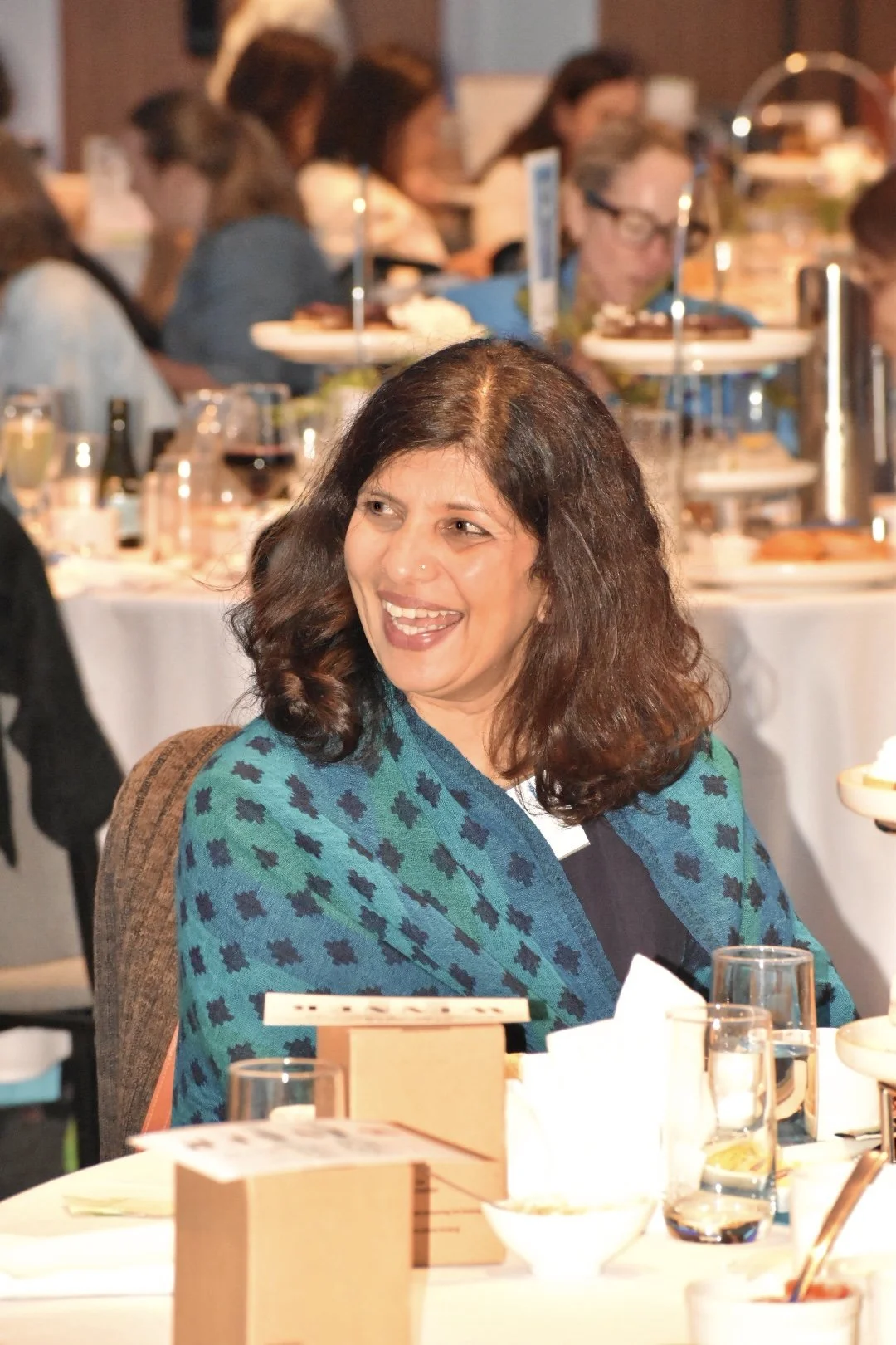 A woman with dark hair smiling at a dinner event, wearing a blue and green patterned shawl, with a busy banquet hall in the background.