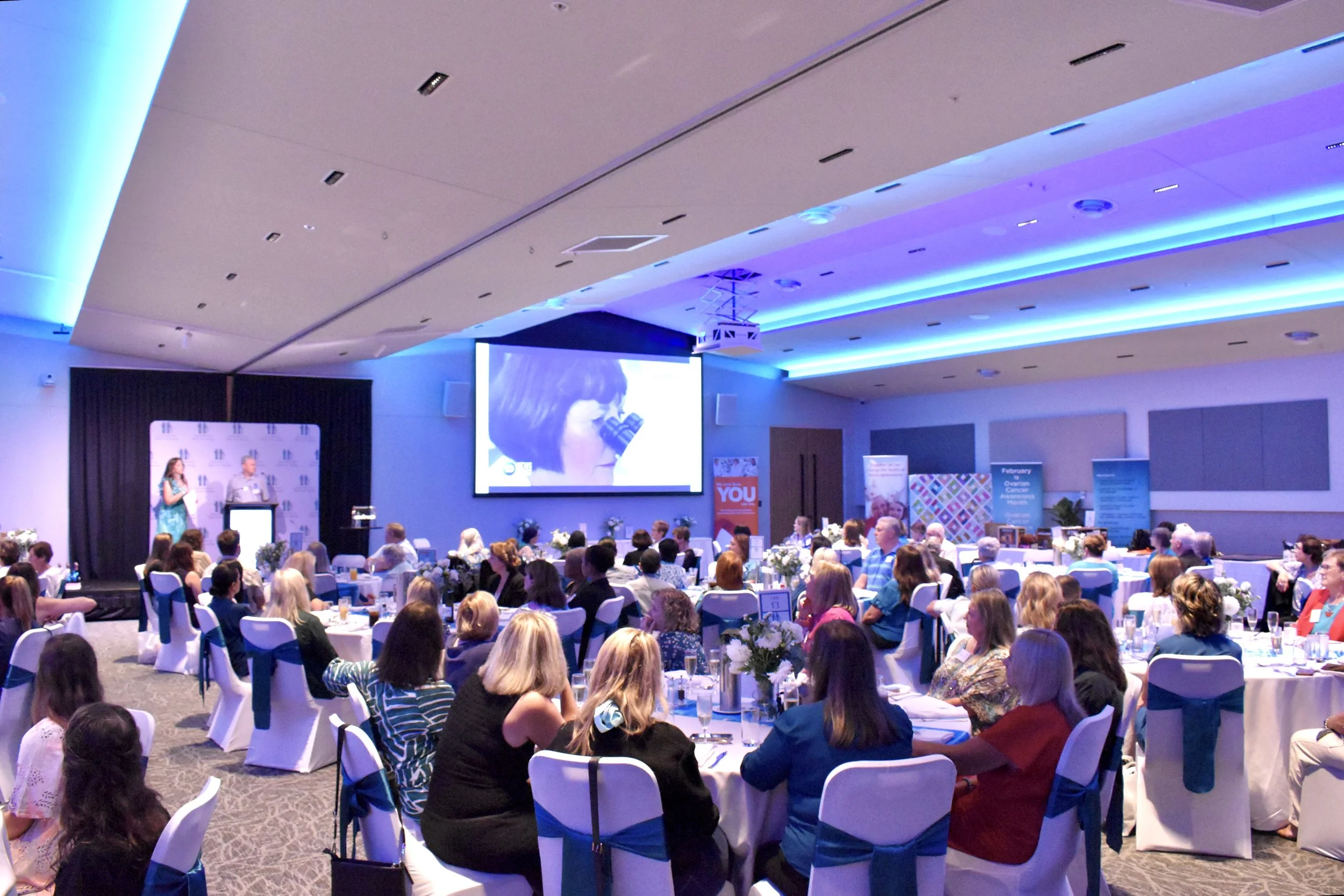 A large conference or seminar room filled with round tables covered in white tablecloths, decorated with floral centerpieces, and filled with mostly women, some men, many wearing professional attire. There is a stage at the front with a podium, and a