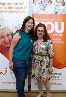 Two women standing together at a health awareness event, smiling. Behind them are banners promoting health and disease prevention, and photos of children wearing medical hats.