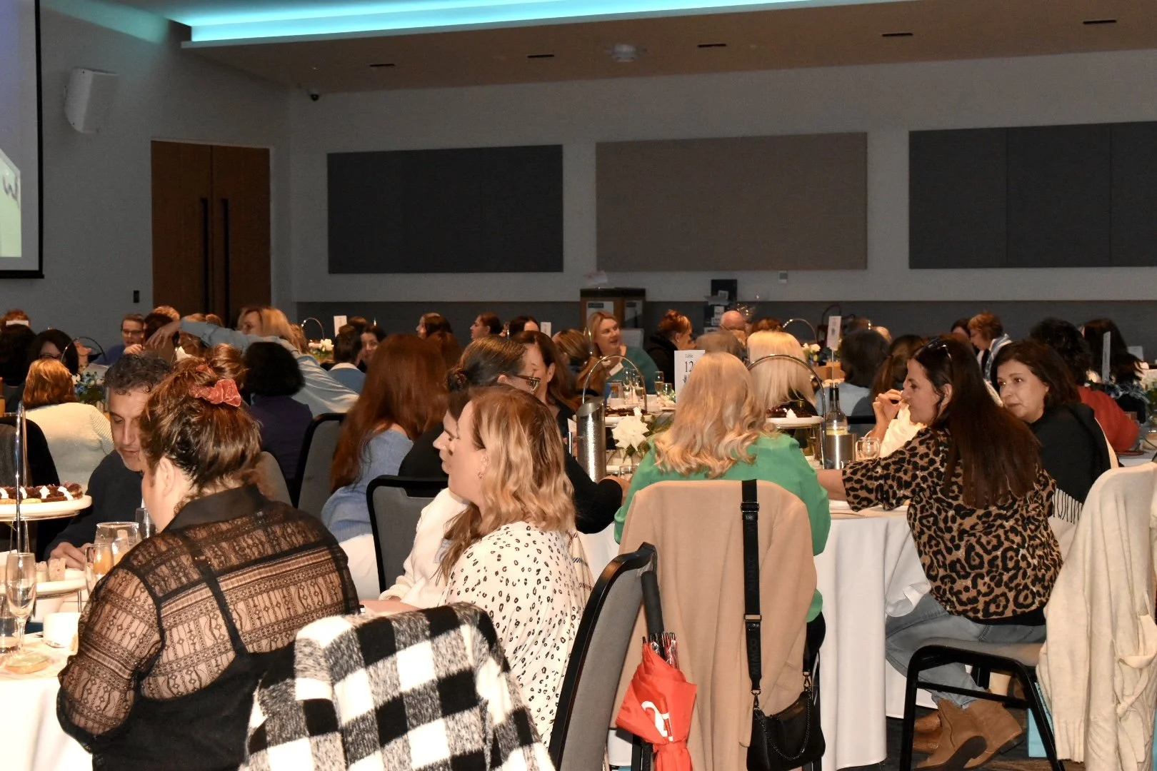 Conference or event with many people seated at round tables in a large room, engaging in conversation and dining.