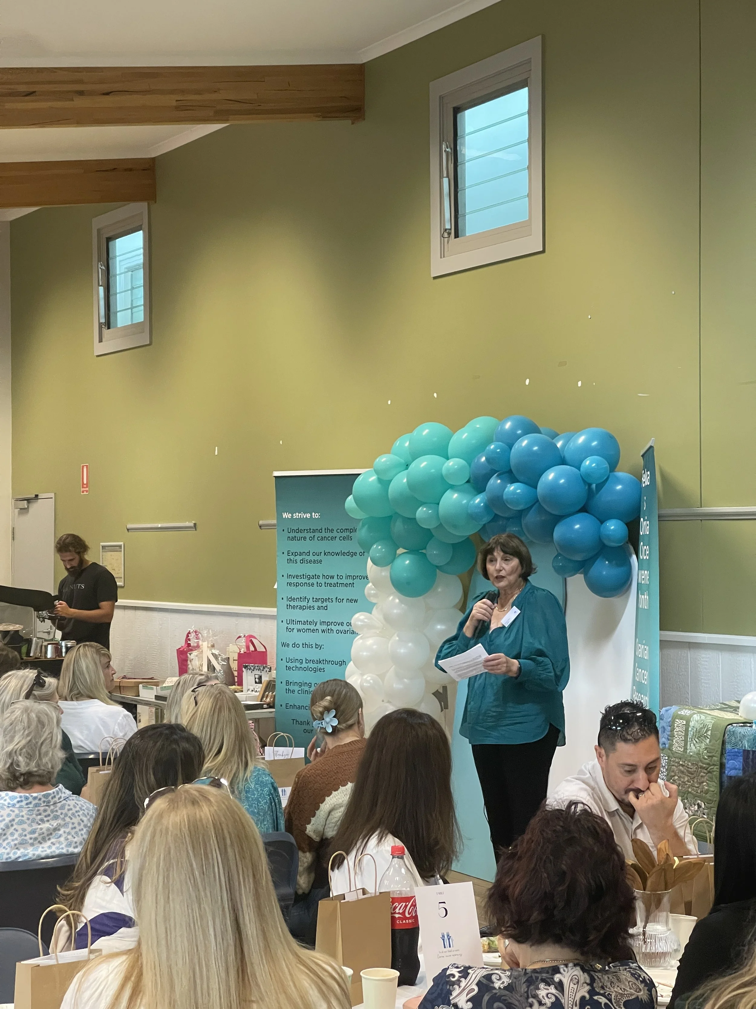 A woman speaking into a microphone at a presentation event, standing next to a balloon arch in an indoor venue, with a banner and seated audience members.