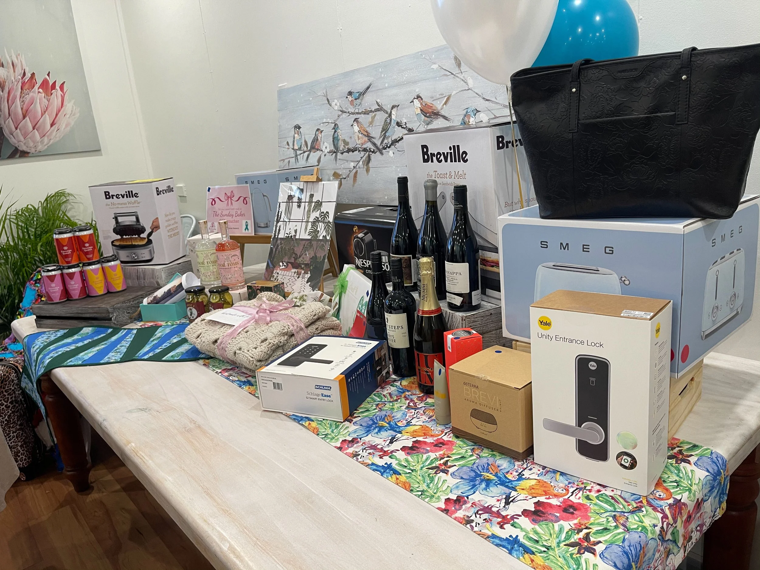 Display of various household and gift items on a table, including wine bottles, a black tote bag, boxes of kitchen appliances, canned goods, bottles of soap, a lock, and decorative items. The table is covered with a colorful floral cloth.