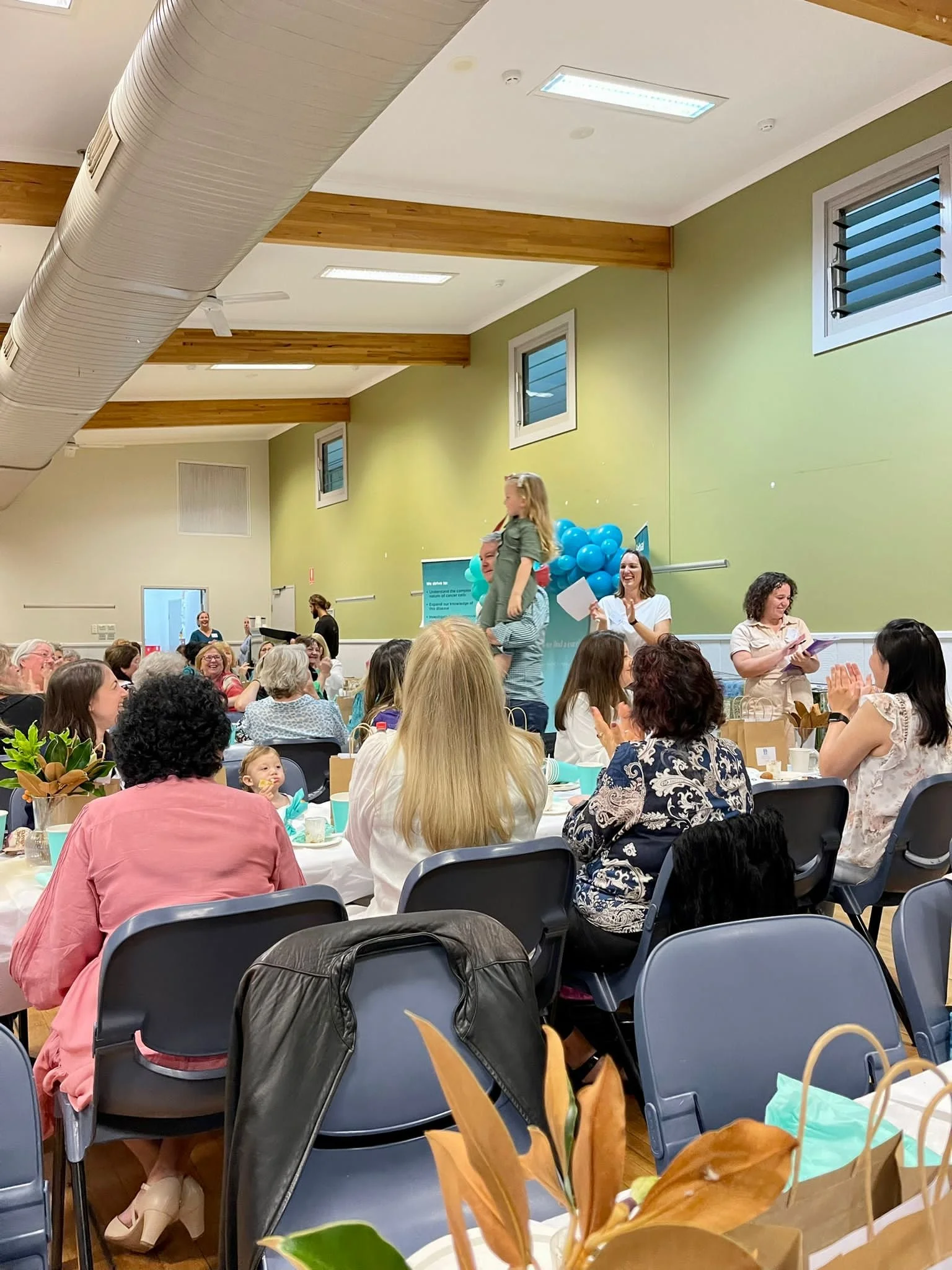 People sitting at tables in a banquet hall, with one young girl standing on another's shoulders on a platform, surrounded by balloons, as part of a celebration or award ceremony.