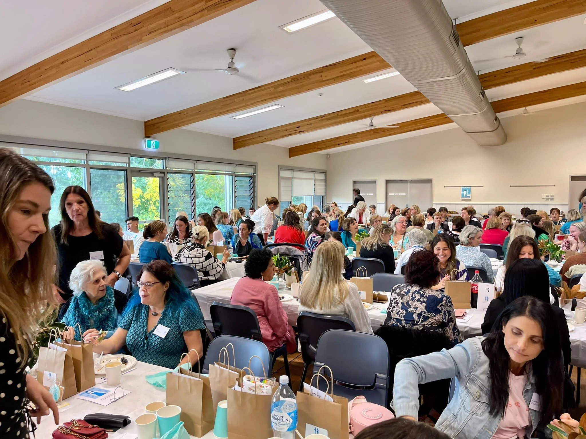 A large indoor gathering with mostly women seated at long tables, engaging in conversation. The room has large windows, wooden beams on the ceiling, and is decorated with table favors and snacks.