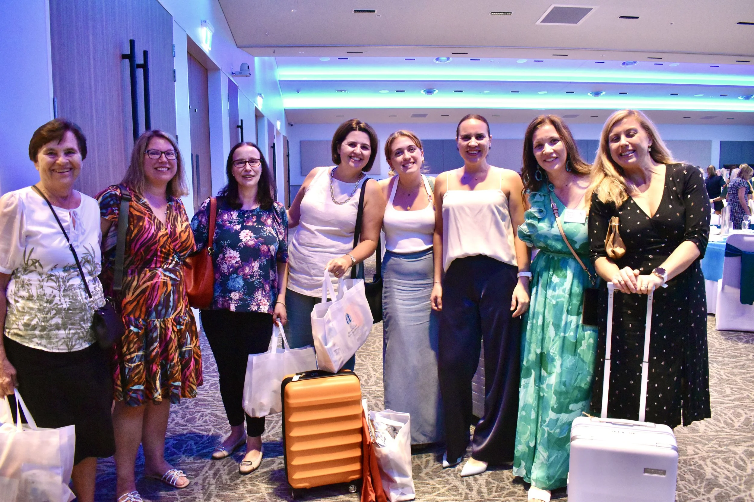 Group of eight women standing together in a conference room, smiling, with luggage and tote bags, in a well-lit event space with round tables and blue lighting.