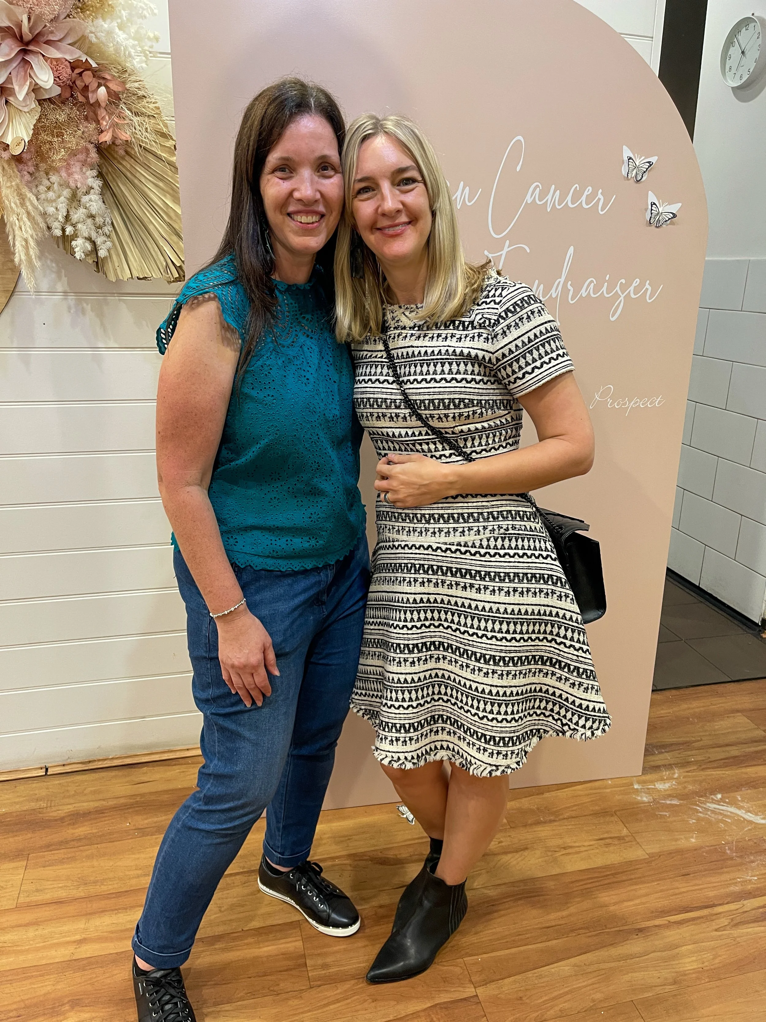 Two smiling women standing close together in front of a backdrop that reads 'Breast Cancer Fundraiser', with decorative flowers and butterflies. One woman is wearing a teal sleeveless top and jeans, and the other is wearing a patterned black and whit