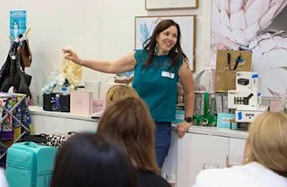 A woman in scrubs standing and smiling in a store or classroom setting, with three people sitting in front, and shelves filled with various items behind her.