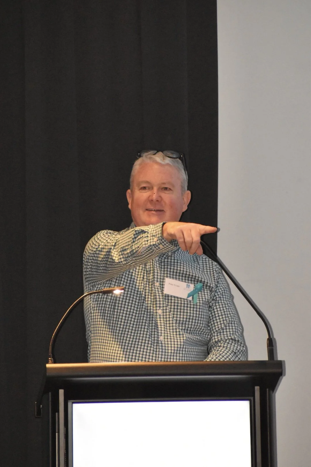 A man standing behind a podium, pointing to the right, wearing a checkered shirt with reading glasses on his head at a conference or event.