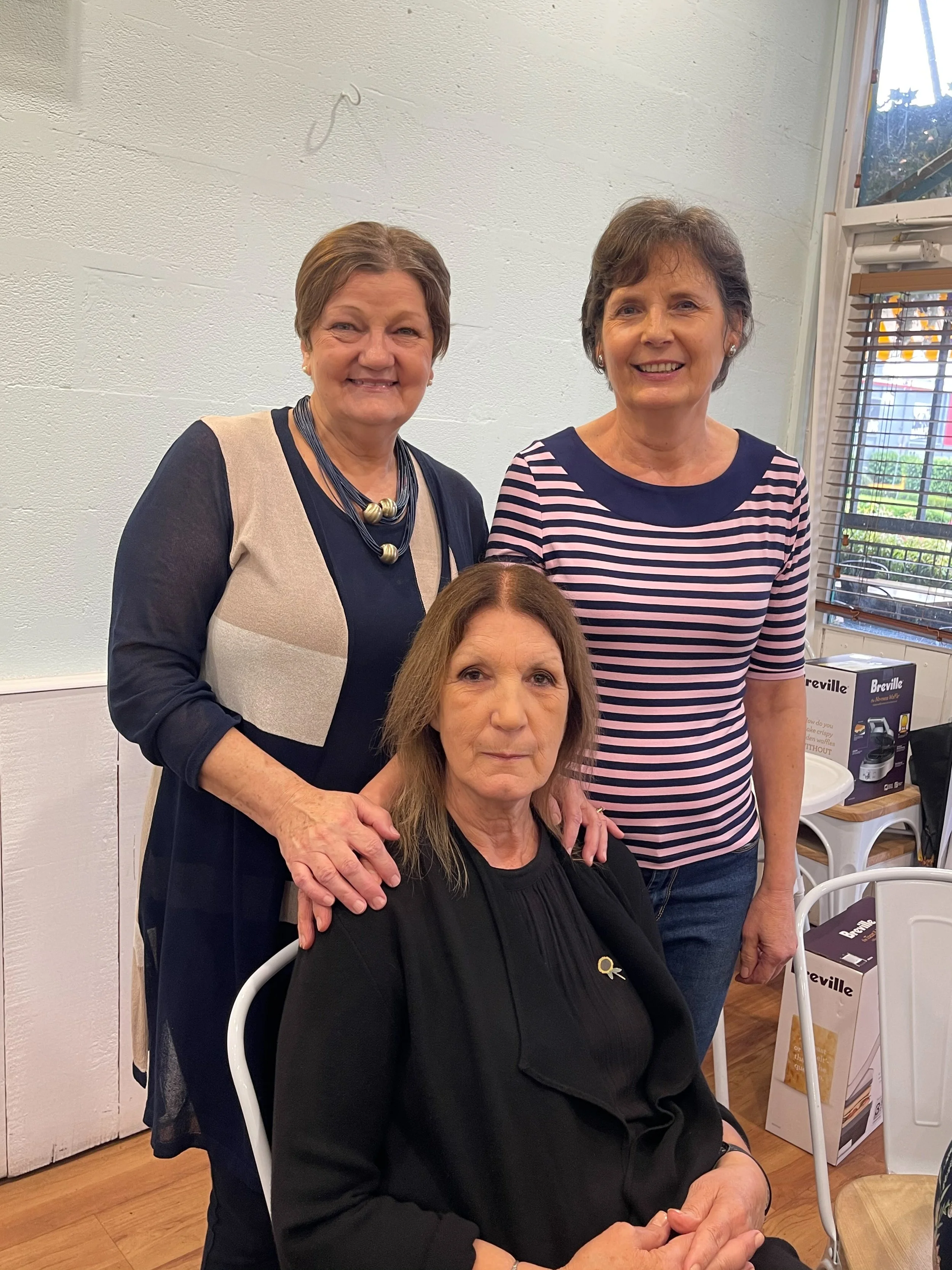 Three women, two standing and one sitting, pose together indoors smiling for the photo. The woman sitting has brown hair and wears a black top. The two women standing have short brown hair; one wears a striped shirt, and the other wears a beige and n