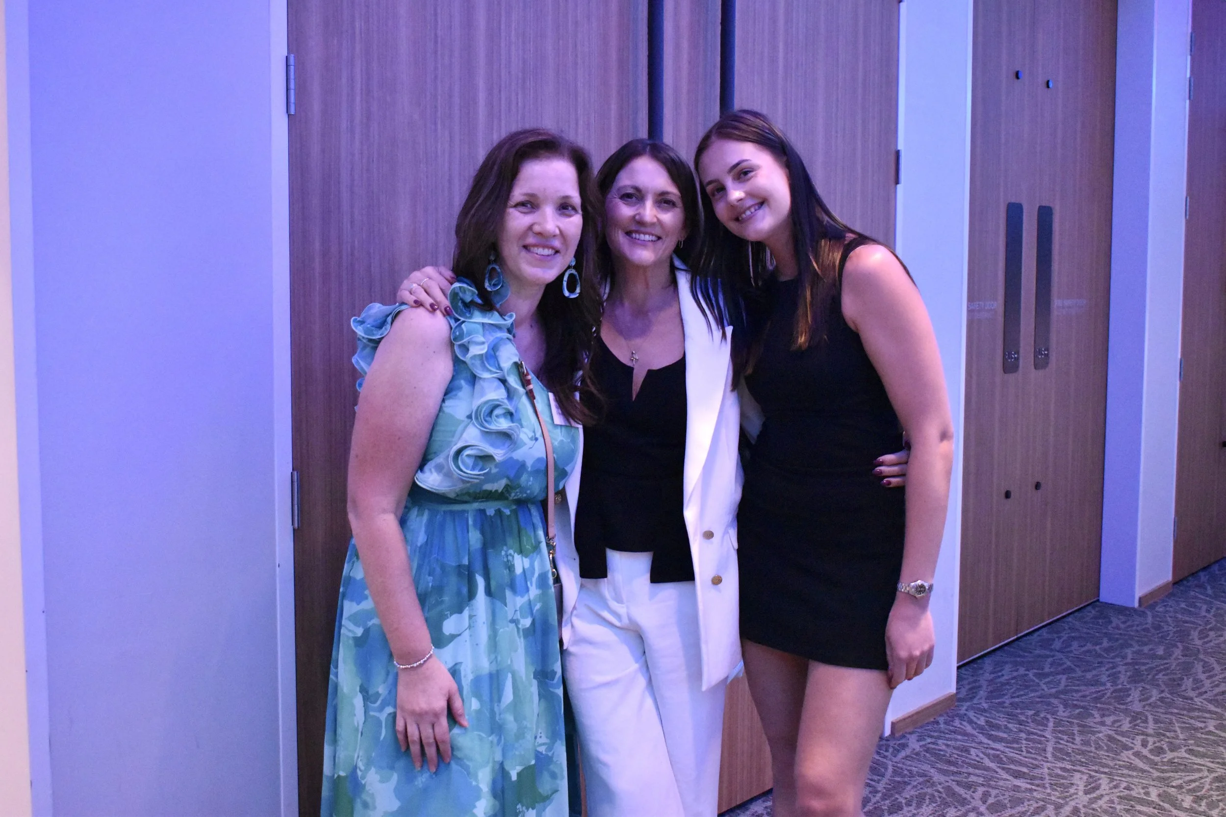 Three women smiling and posing together indoors near wooden doors.